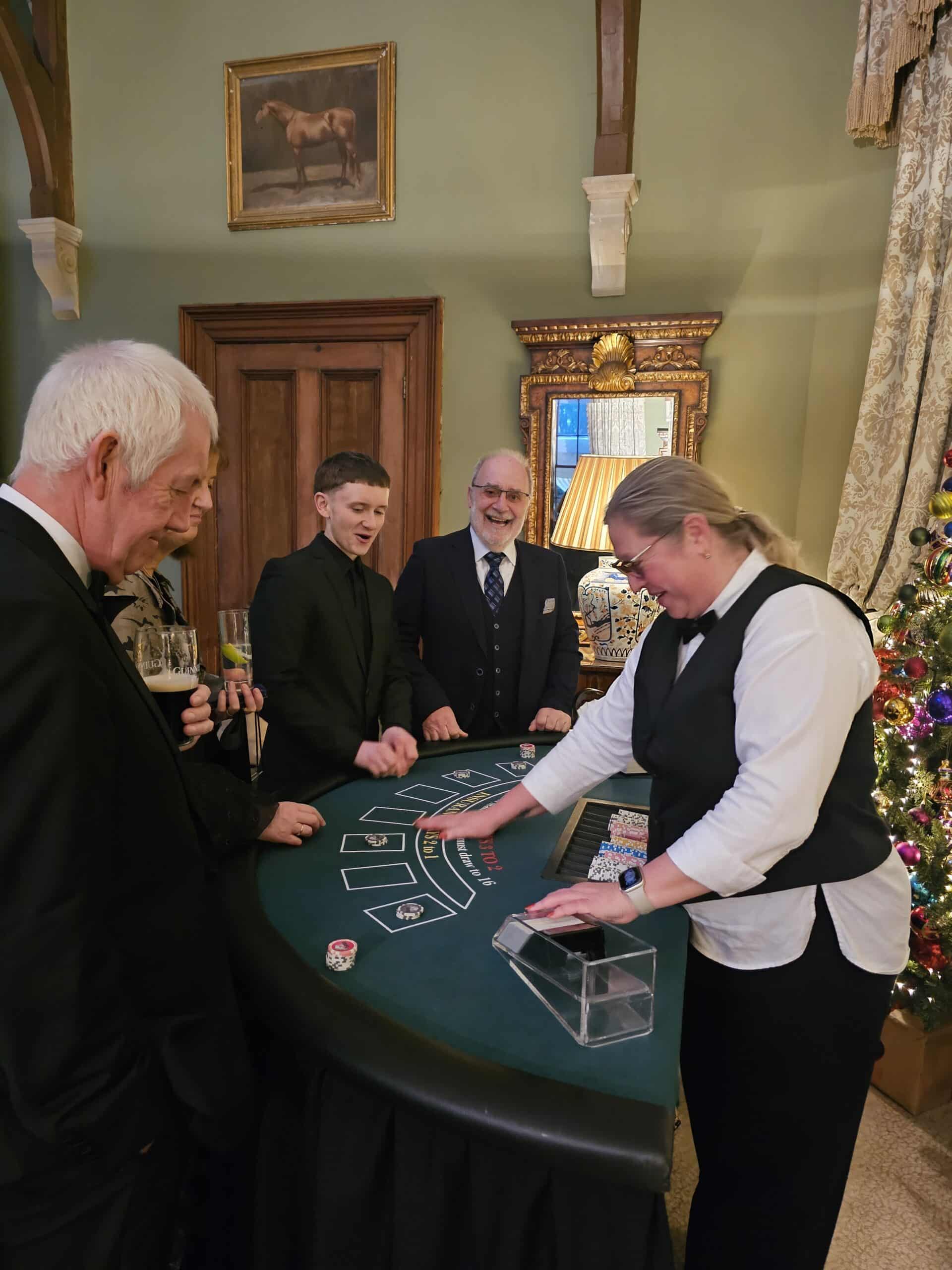 People playing blackjack at a green table, bartender dealing cards, festive room with decorated Christmas tree in background.