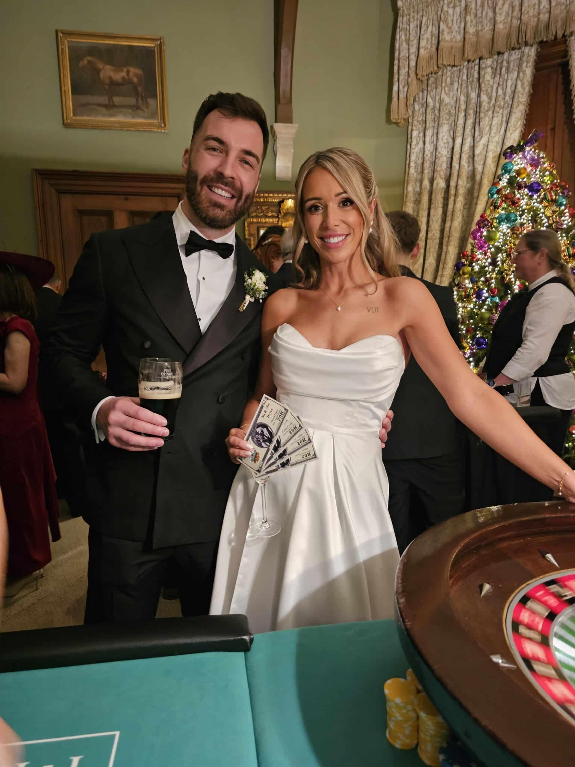 A couple dressed formally at a casino-themed event, holding a drink and play money near a roulette table with a decorated Christmas tree in the background.