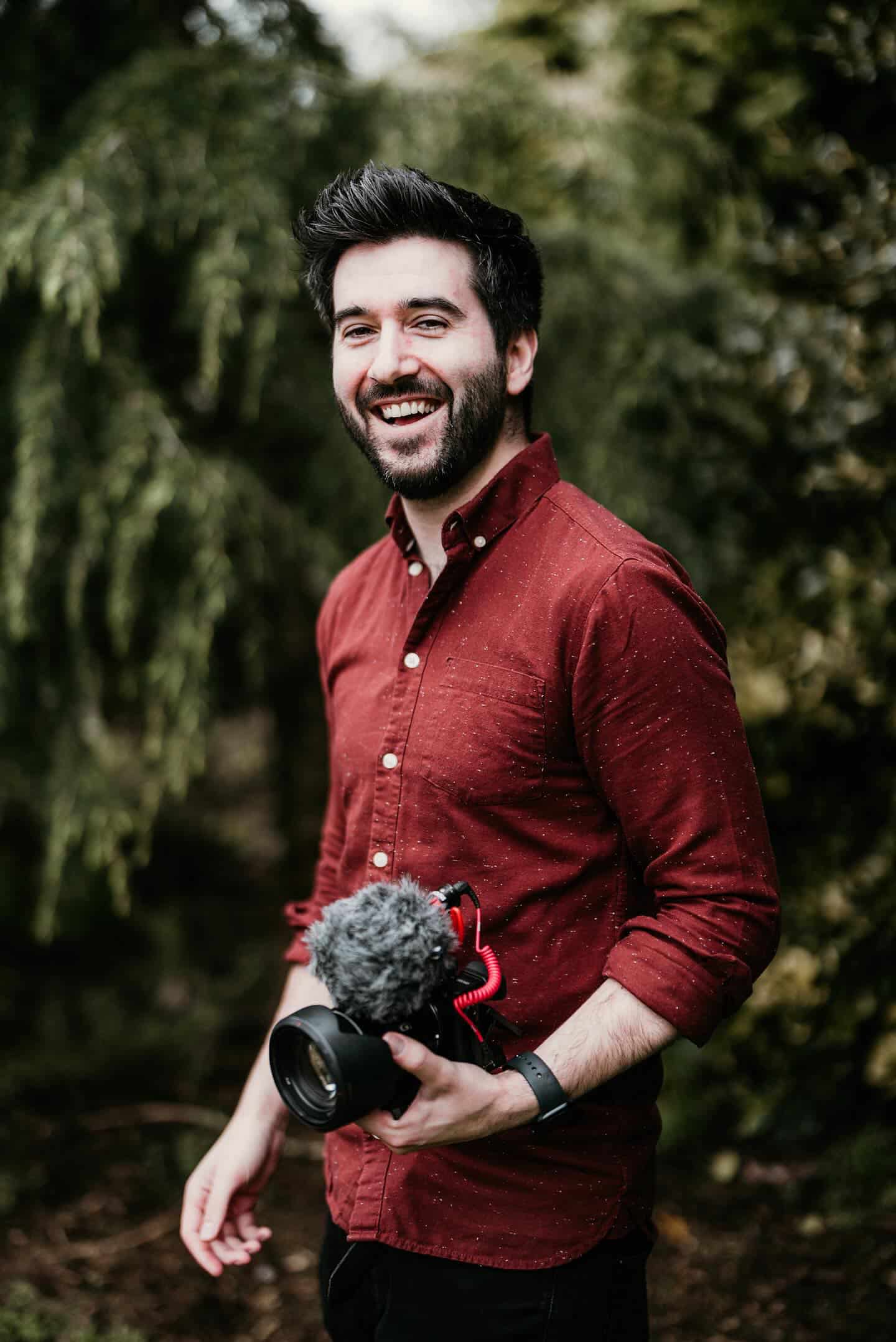 Smiling man in a red shirt holding a camera with a microphone, standing outdoors against a blurred green background.
