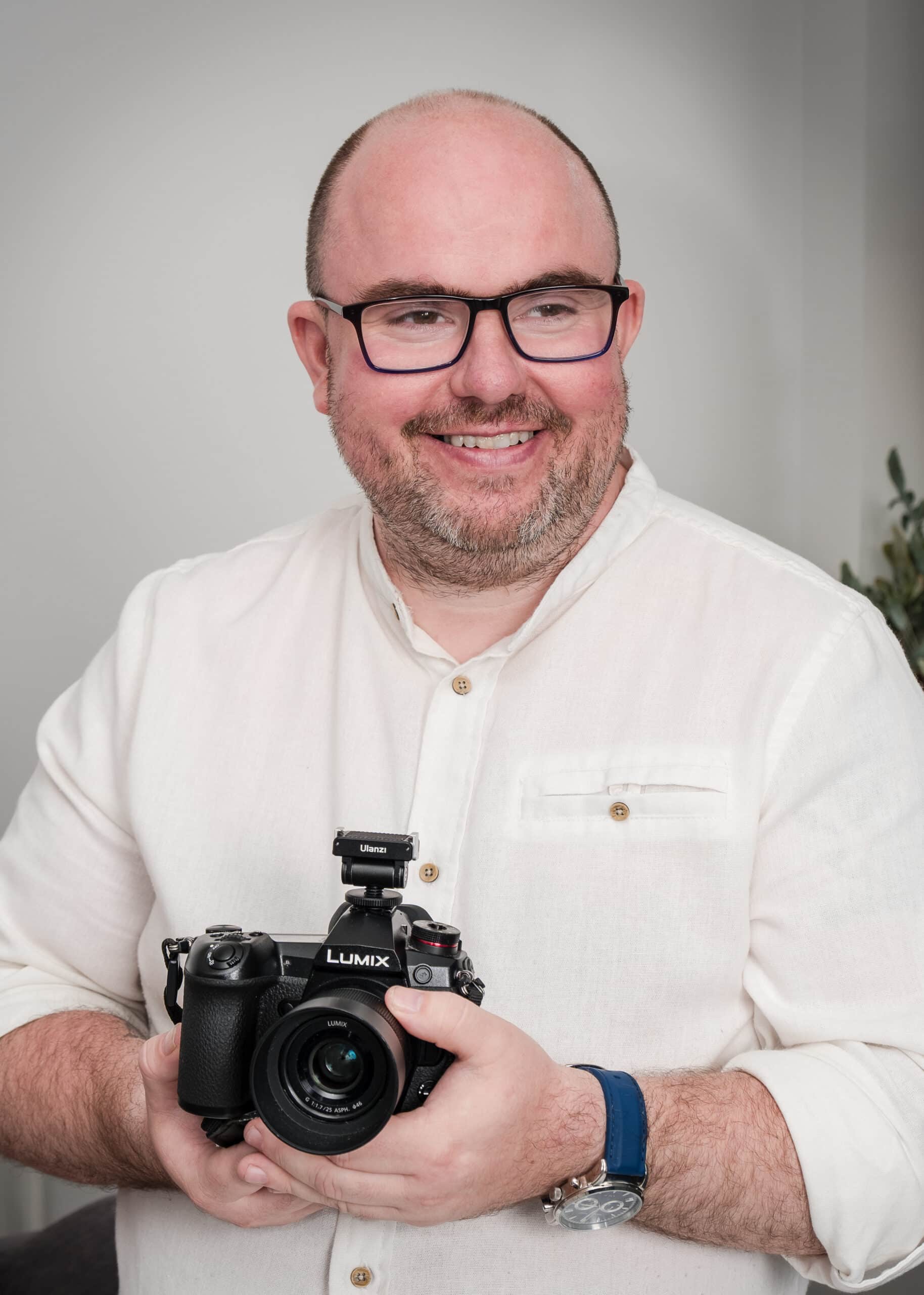 Man holding a Lumix camera, wearing glasses and a white shirt, smiling.