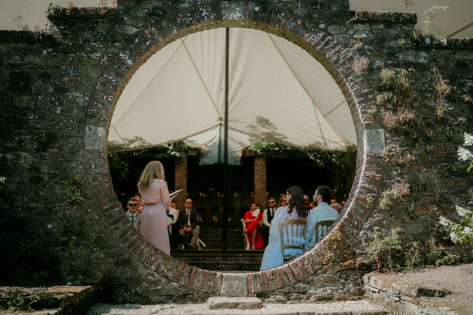 Wedding ceremony with guests seated in a garden setting, viewed through a circular stone arch.