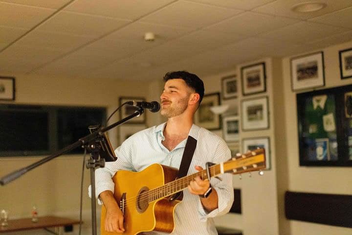 Musician singing and playing an acoustic guitar in a room with framed pictures on the walls.