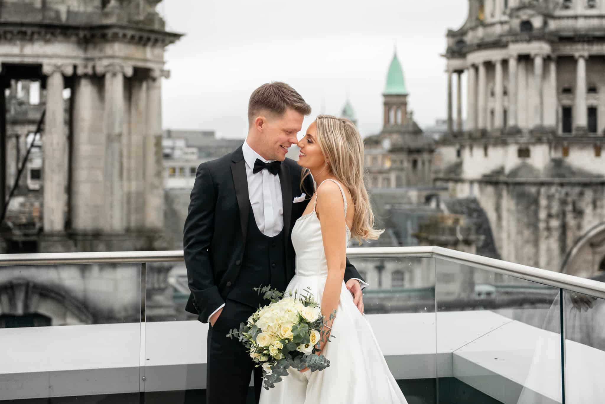 Bride and groom embracing on a rooftop with architectural city backdrop.