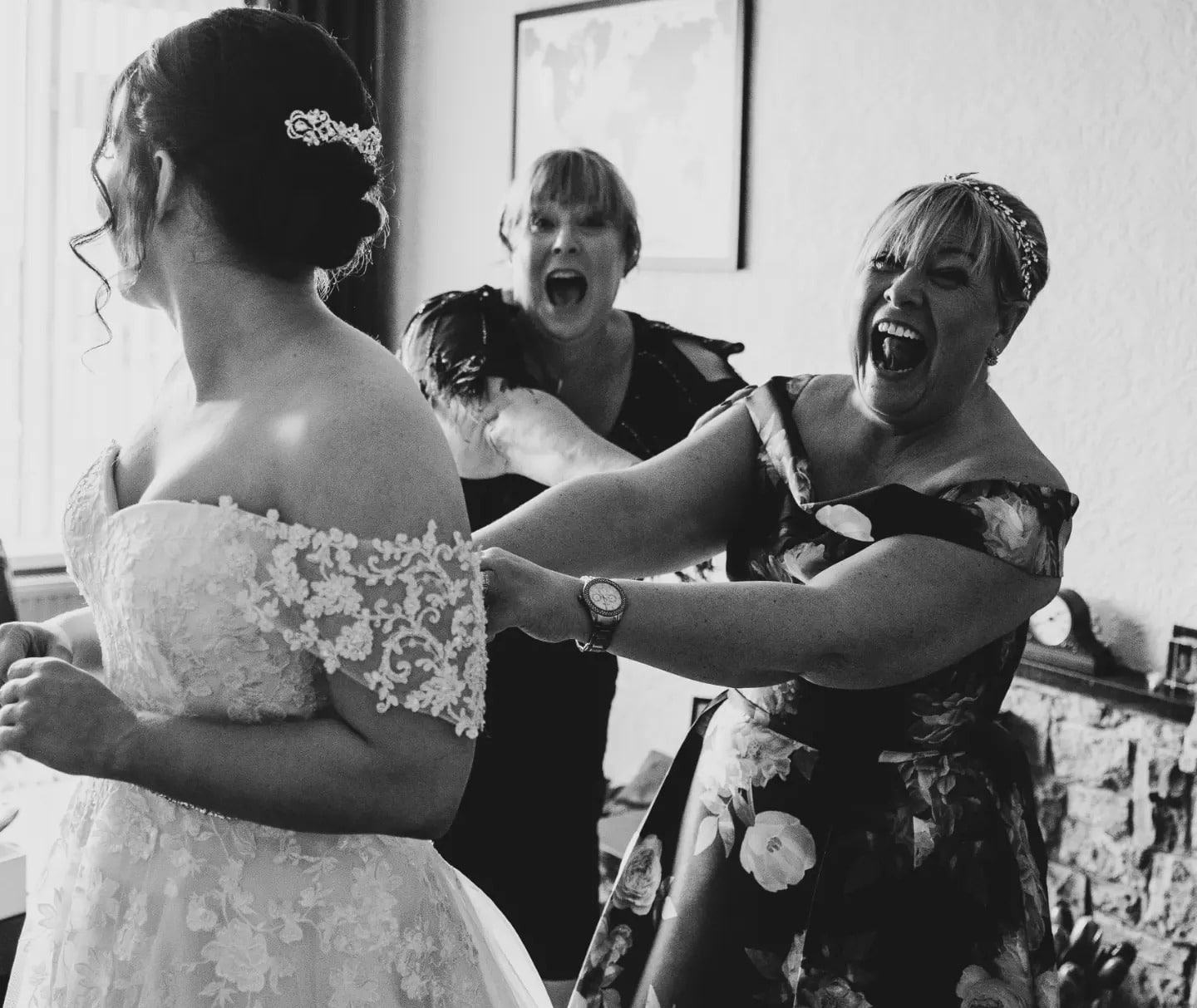 Three women joyfully assist a bride with her wedding gown in a lively, candid moment.