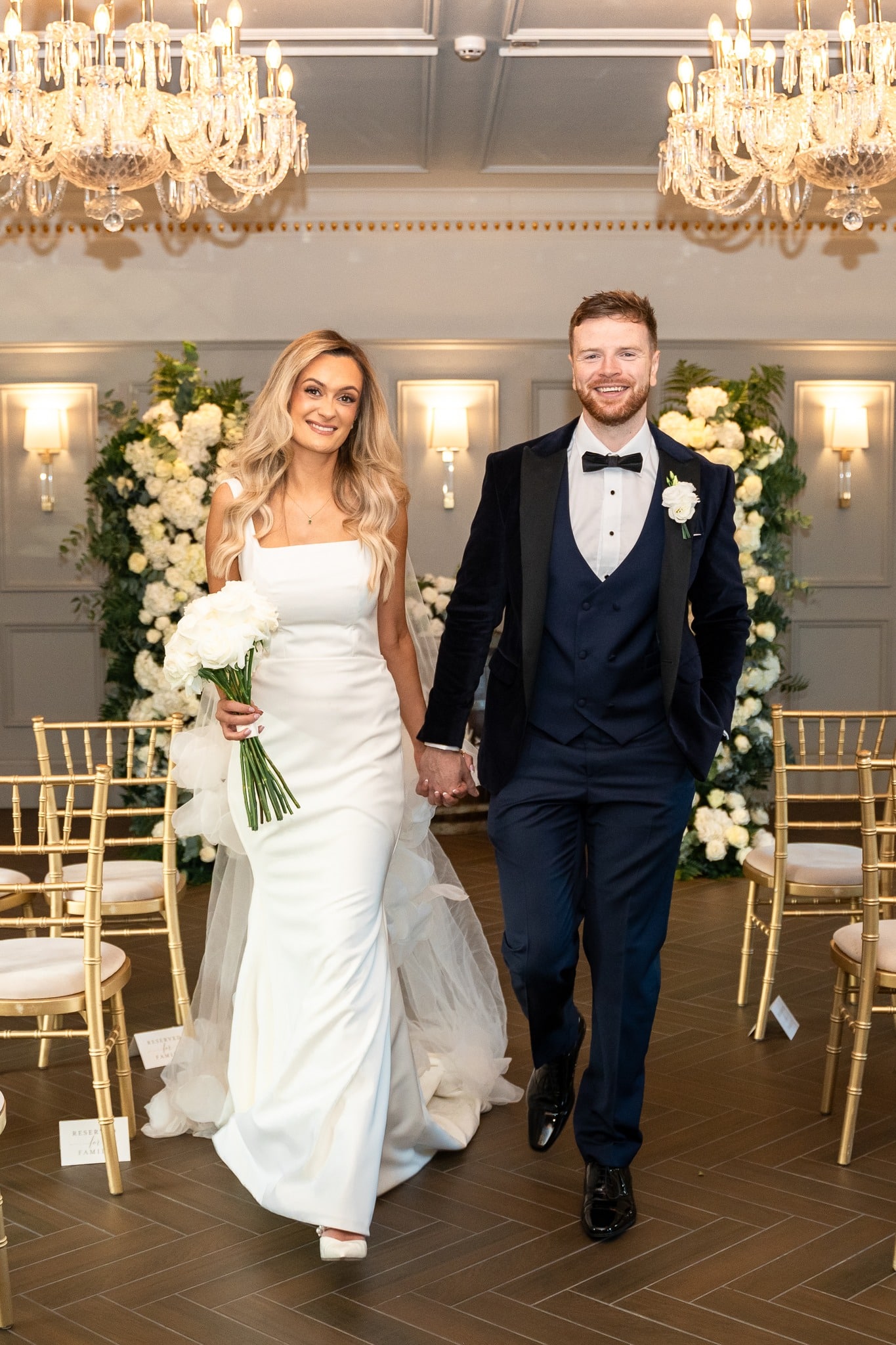 A bride and groom joyfully walking down the aisle in an elegant wedding venue with floral decor and chandeliers.