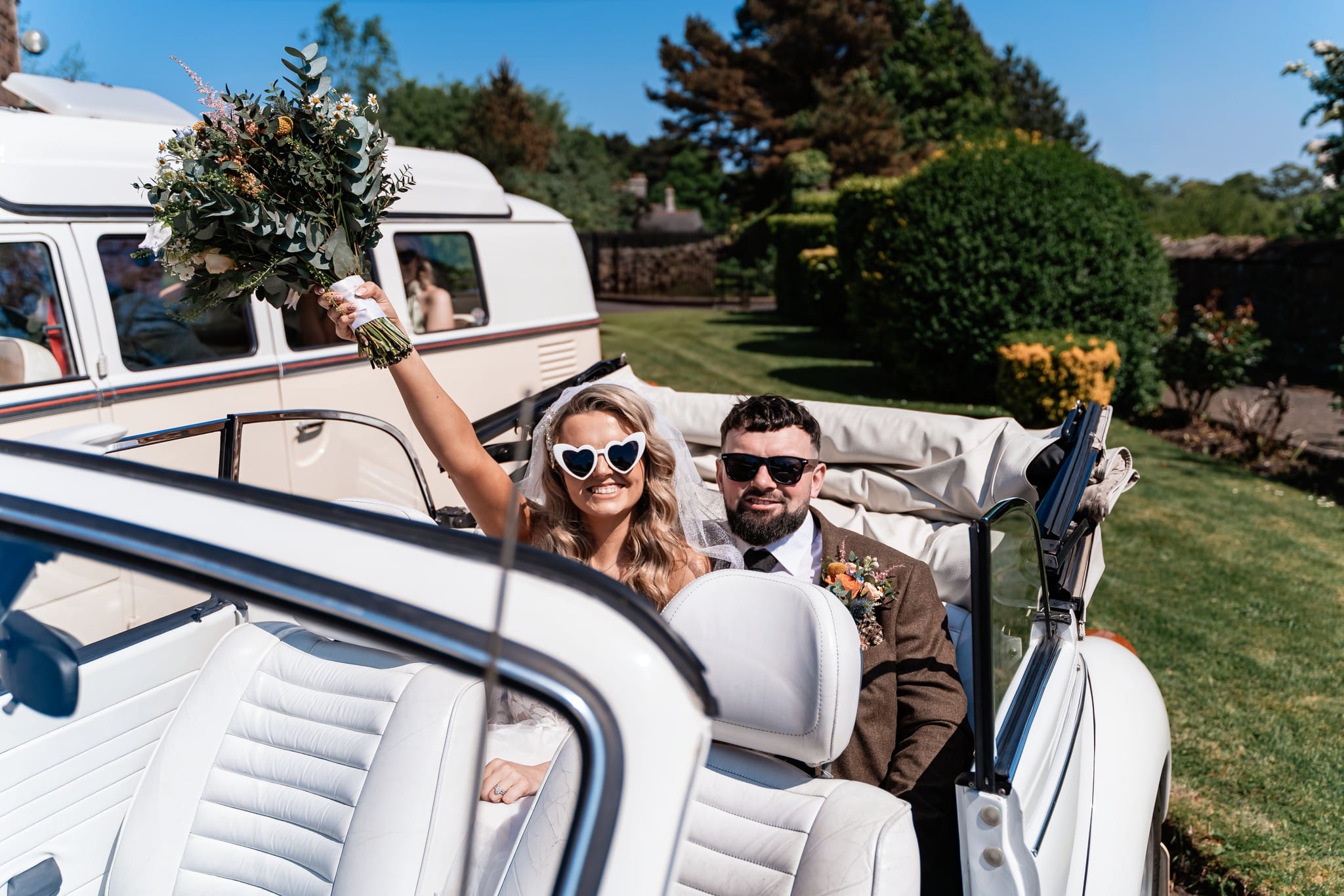 Bride and groom in convertible car, bride holding bouquet and wearing heart-shaped sunglasses.