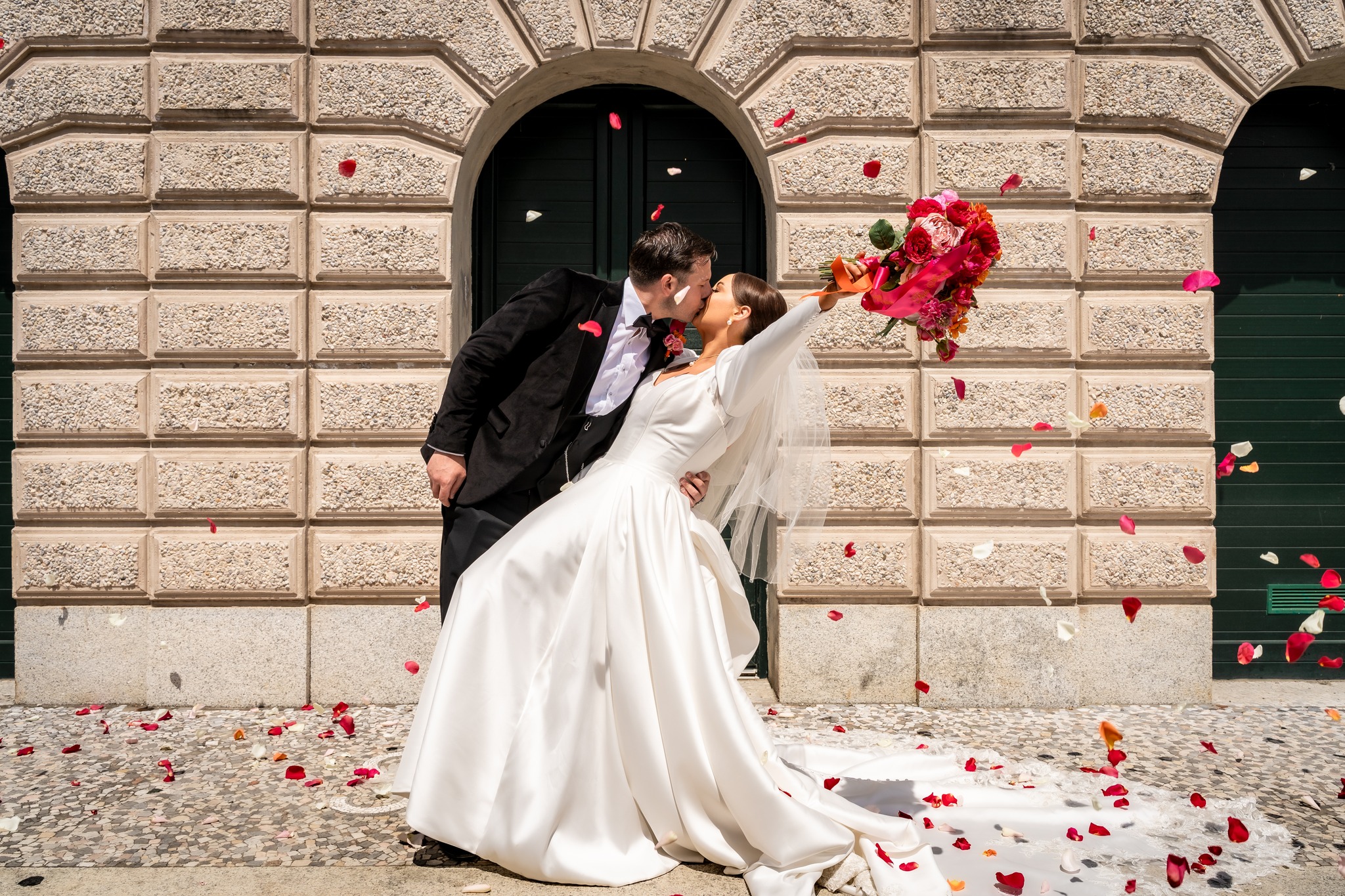 Bride and groom kissing, bride holding bouquet, rose petals falling, outside brick building.