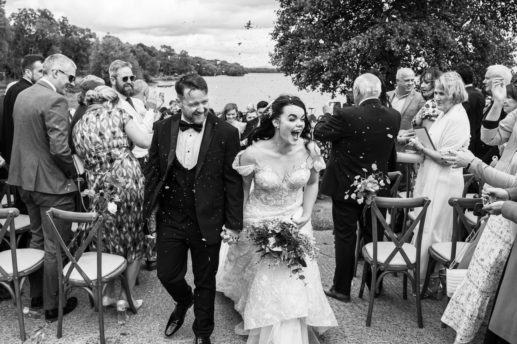 Bride and groom joyfully walking down aisle with guests throwing confetti outdoors.