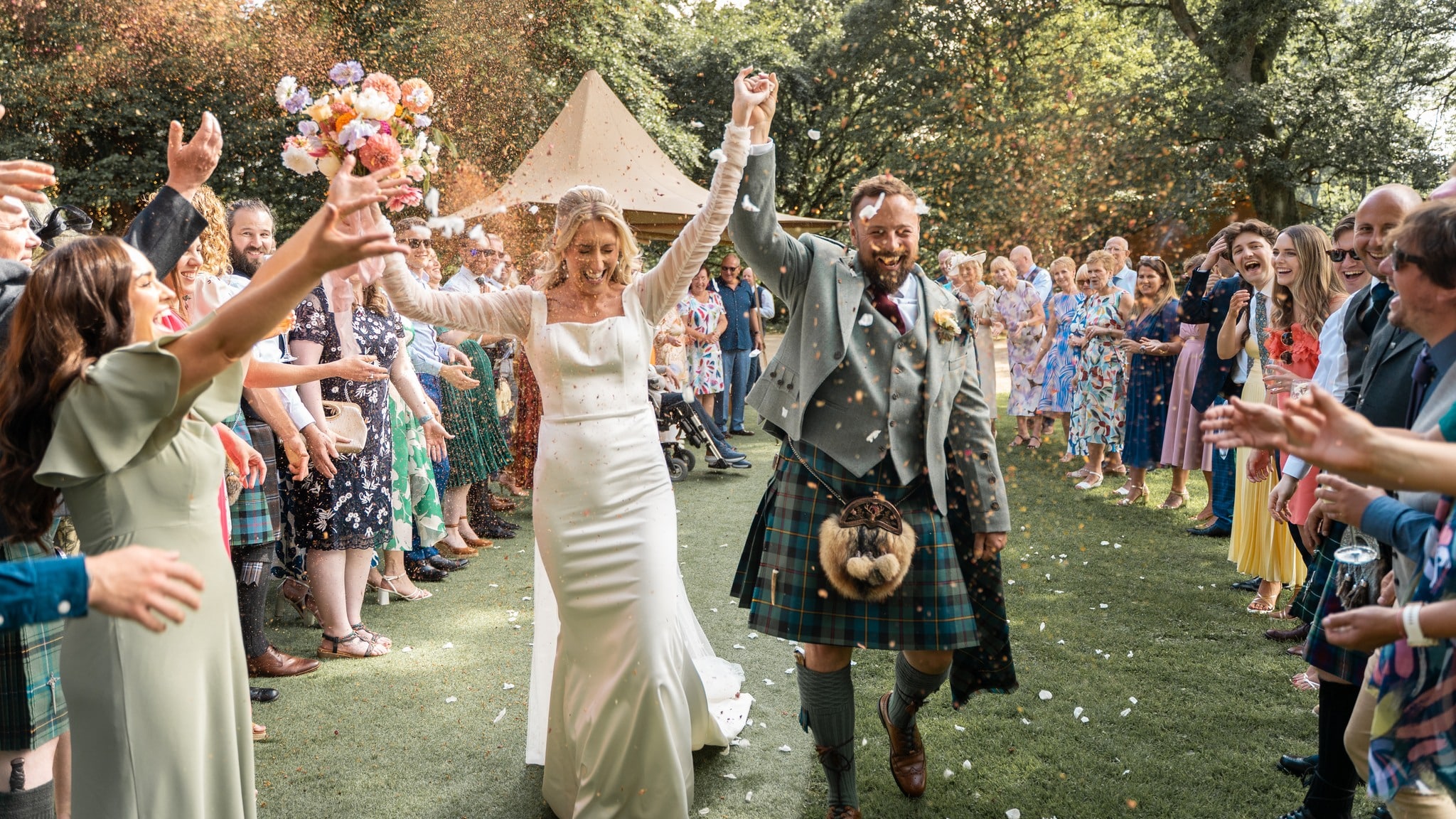 Bride and groom joyfully walk through a cheering crowd outdoors, while colorful confetti fills the air.