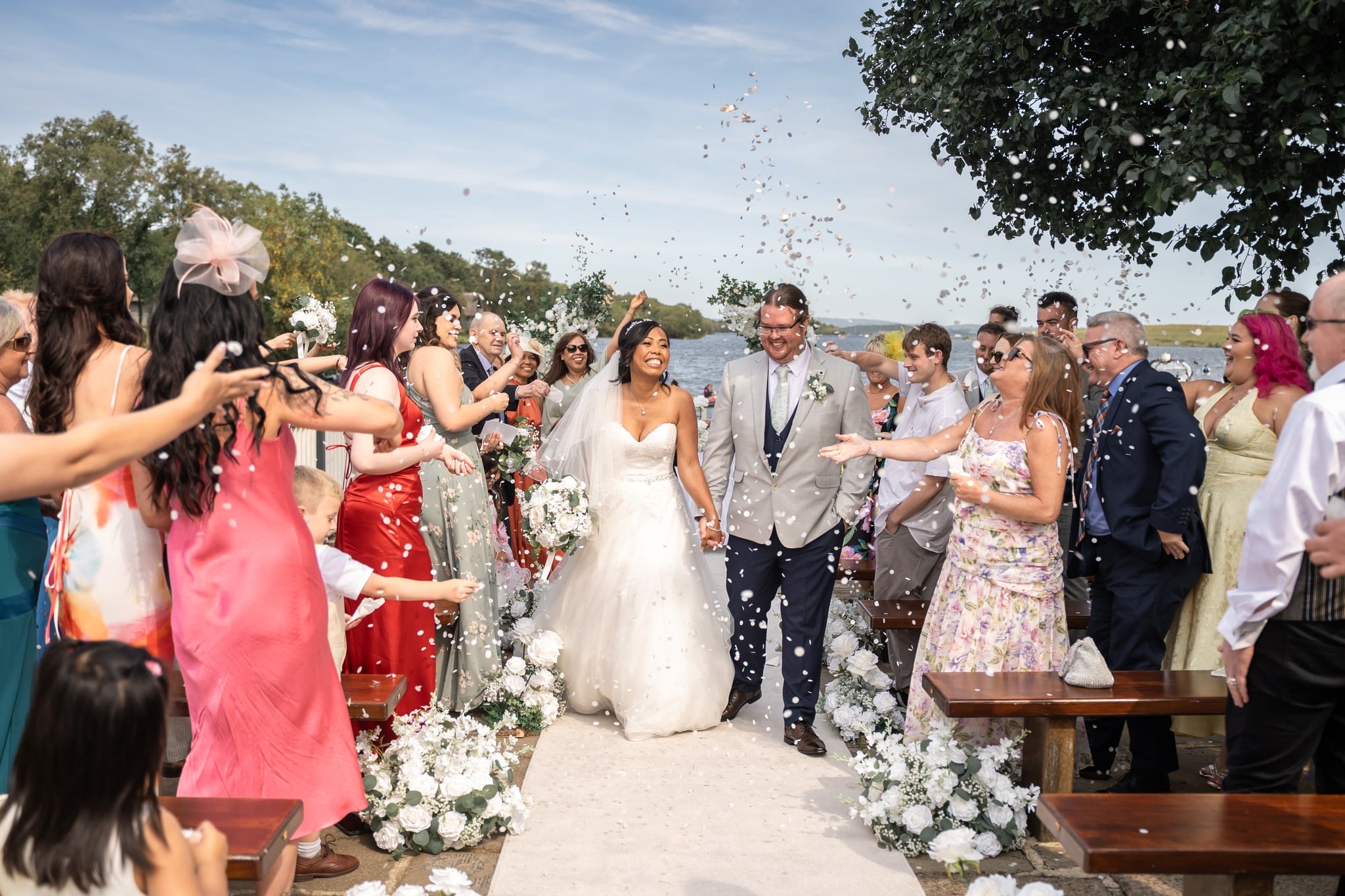 Bride and groom walk through a joyful crowd throwing confetti at an outdoor wedding ceremony.
