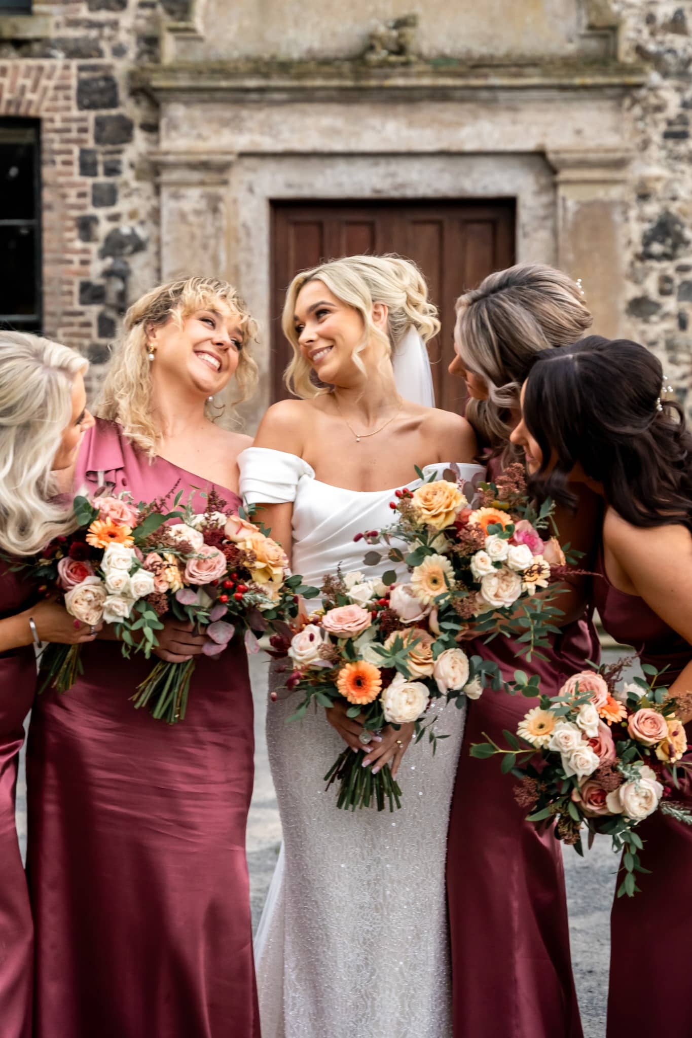 Bride with bridesmaids in burgundy dresses holding bouquets, smiling joyfully outside a stone building.