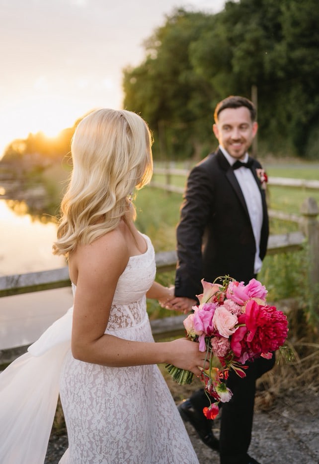 Bride and groom holding hands at sunset, bride holding a bouquet of pink and red flowers.