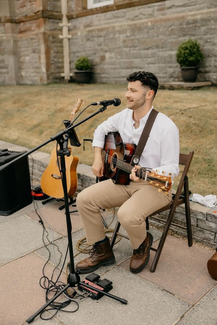 Man playing acoustic guitar and singing outdoors with a microphone and equipment setup.