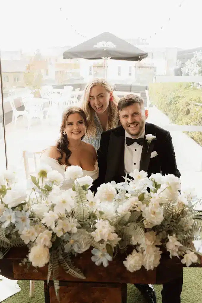 Bride and groom with a guest pose behind a floral arrangement in an outdoor setting.