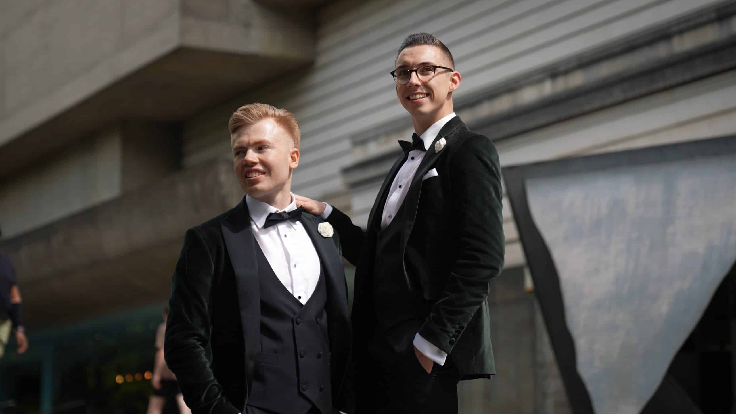 Two people in formal suits smiling, standing in an outdoor urban setting.