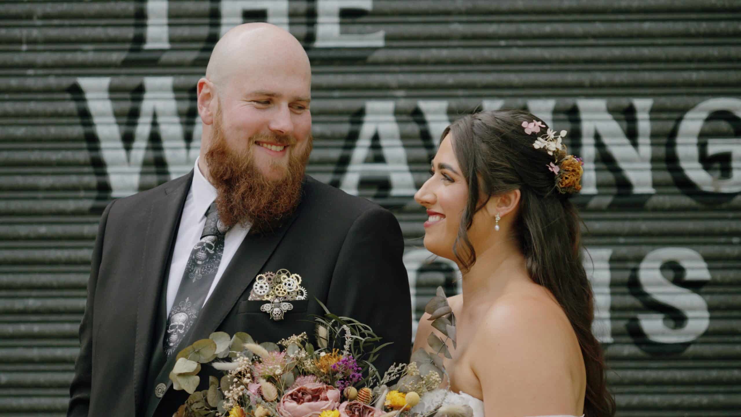Smiling couple in wedding attire with bride holding a floral bouquet, standing against a backdrop with text.