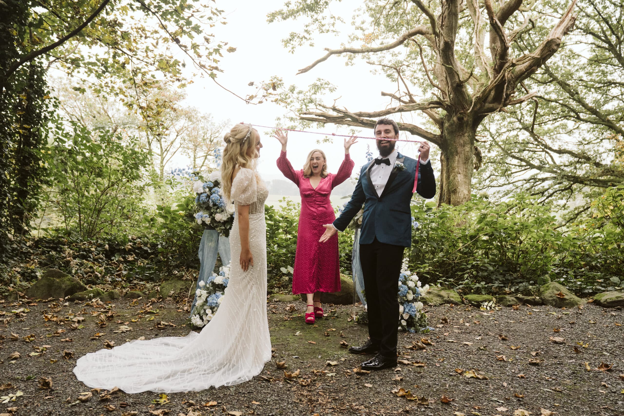 Bride and groom at outdoor wedding ceremony with officiant, surrounded by trees and floral decorations.