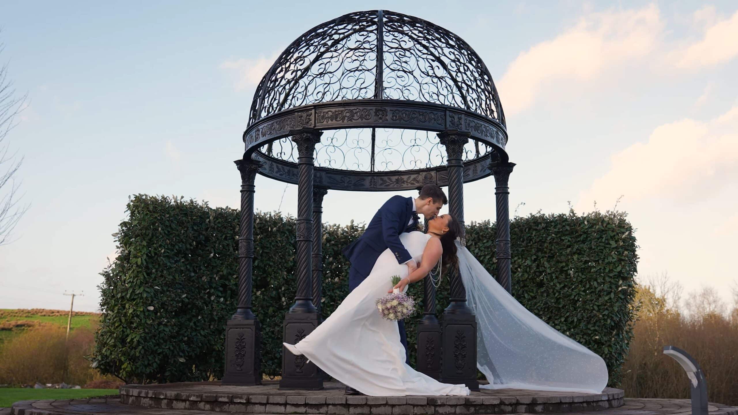 Bride and groom kiss under ornate metal gazebo with greenery backdrop.