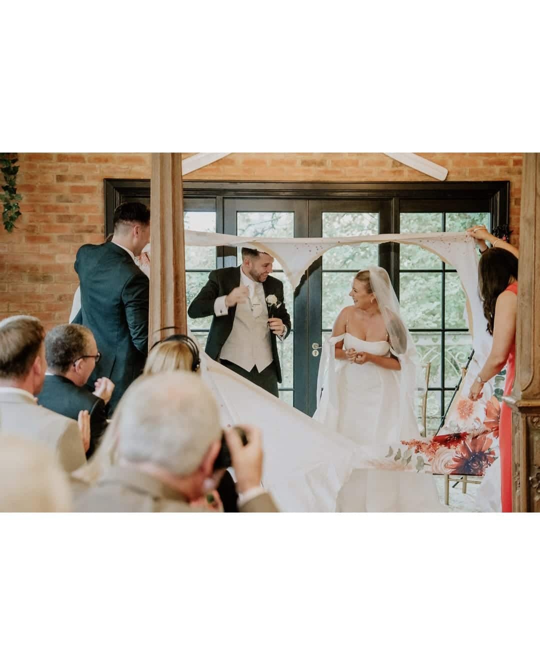 Bride and groom laughing while holding a cut-out heart banner, surrounded by guests.