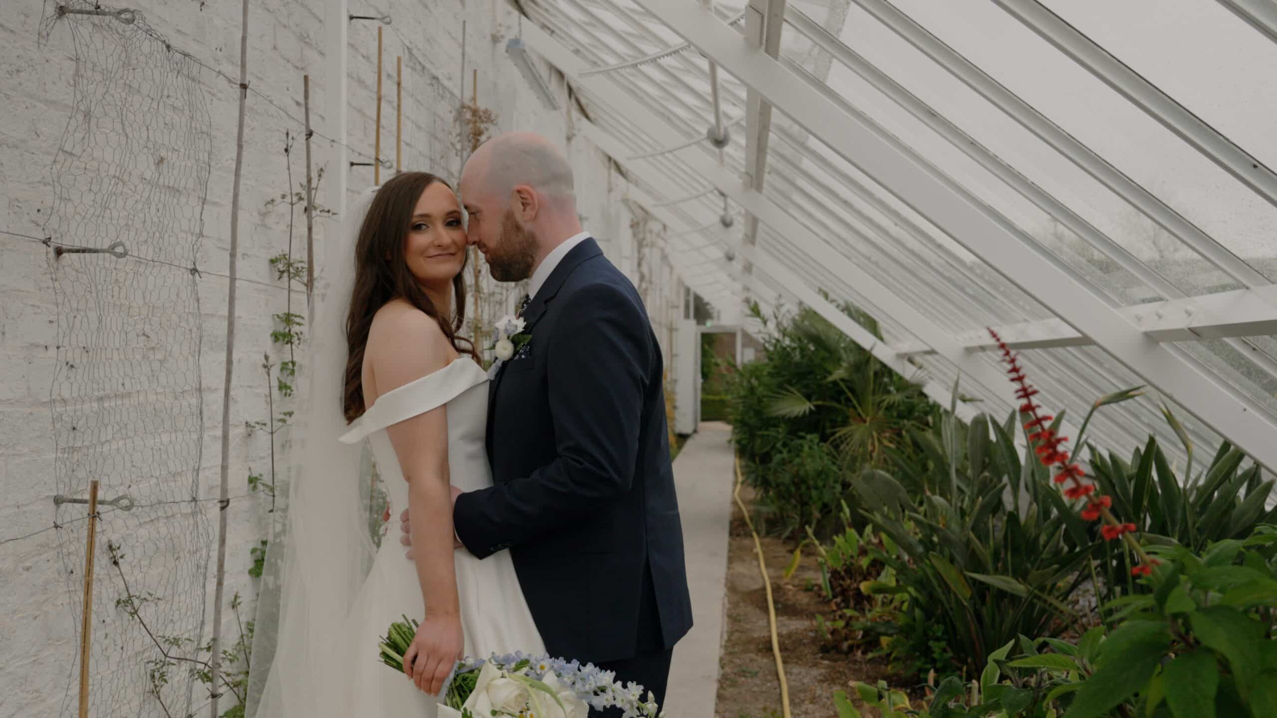 Bride in white gown and groom in suit embracing in a greenhouse, surrounded by greenery.