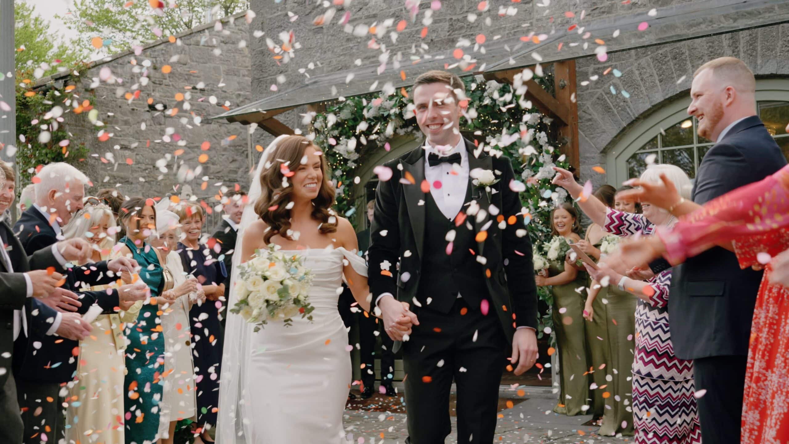Bride and groom walking through falling confetti at wedding ceremony, surrounded by guests.