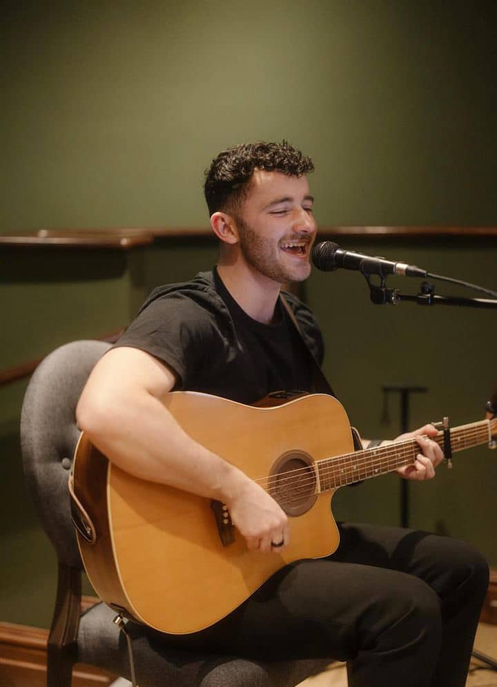 A man playing an acoustic guitar and singing into a microphone in a cozy indoor setting.