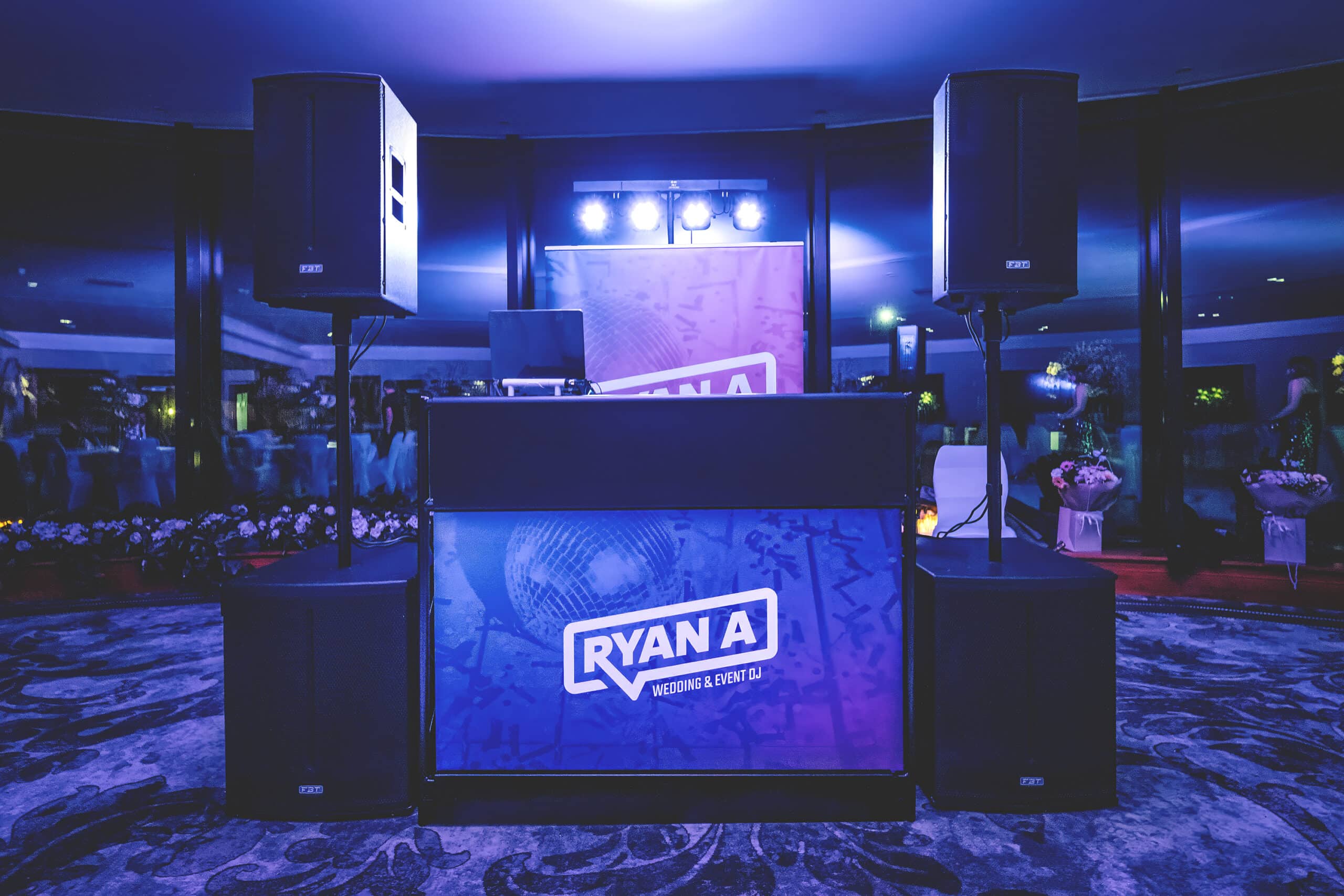 DJ setup with speakers and laptop in a dimly lit venue, decorated with vibrant lighting and floral arrangements.