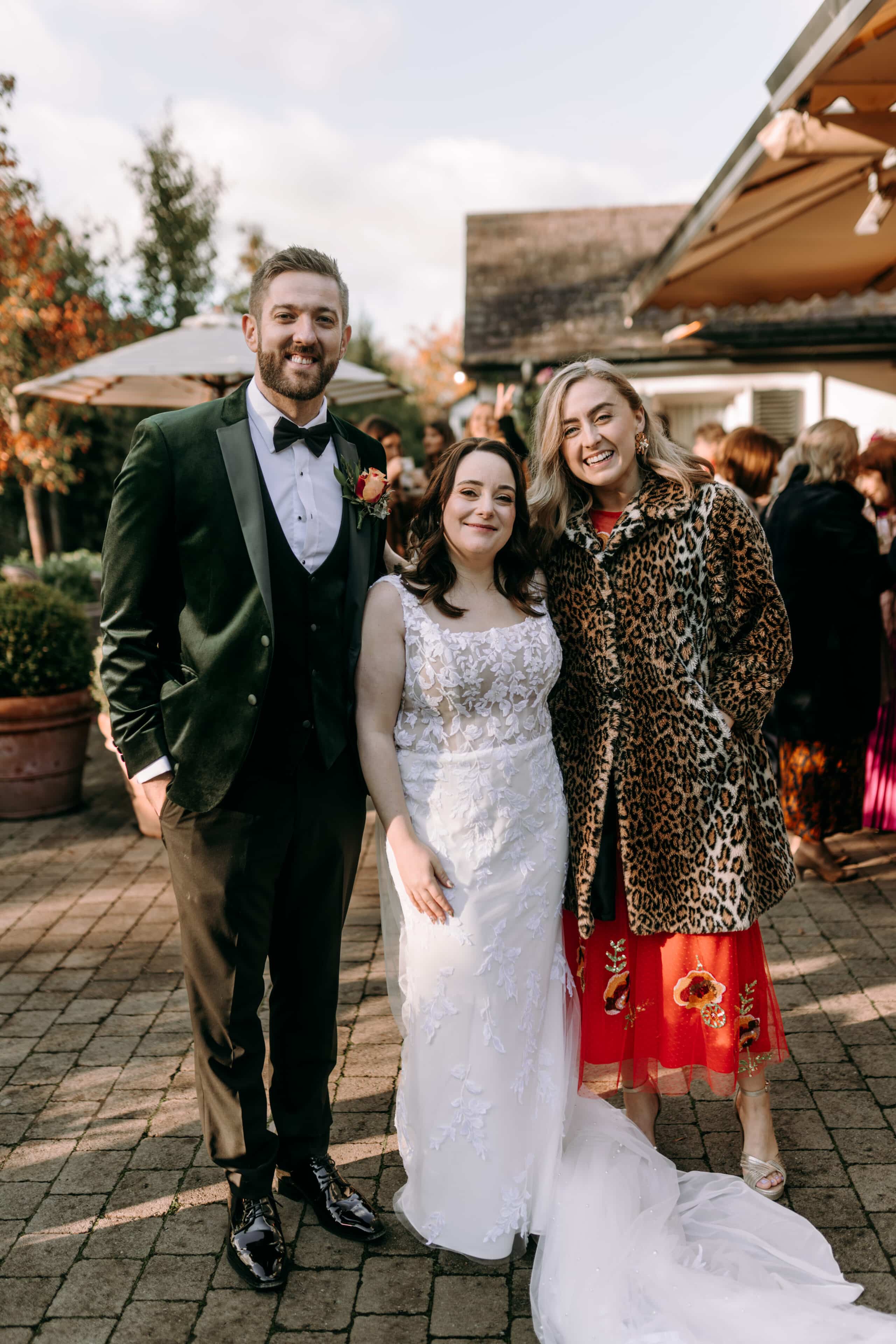 Bride and groom standing outdoors with a guest in a leopard print coat, surrounded by wedding attendees.