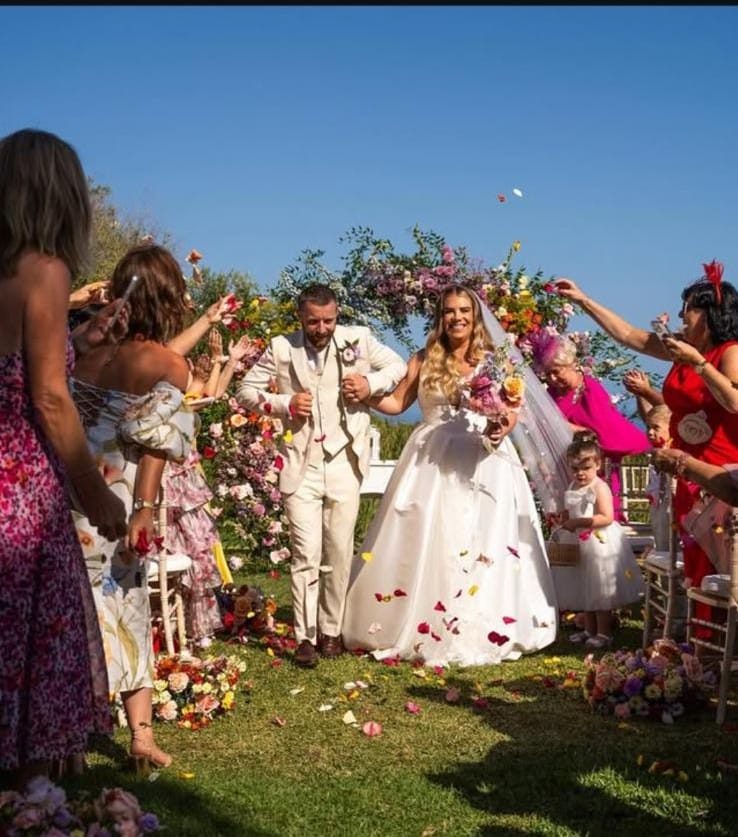 Bride and groom walking down the aisle, surrounded by guests, flowers, and confetti in a garden wedding ceremony.
