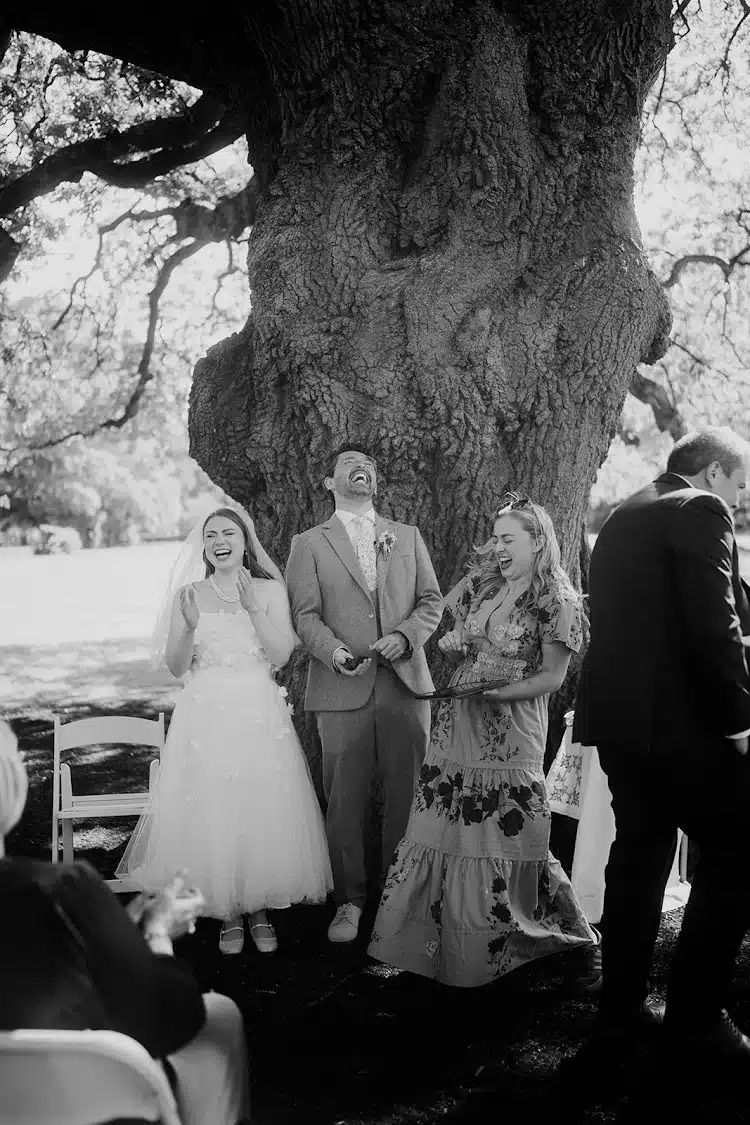 Bride, groom, and guest laughing joyfully in an outdoor wedding ceremony under a large tree.