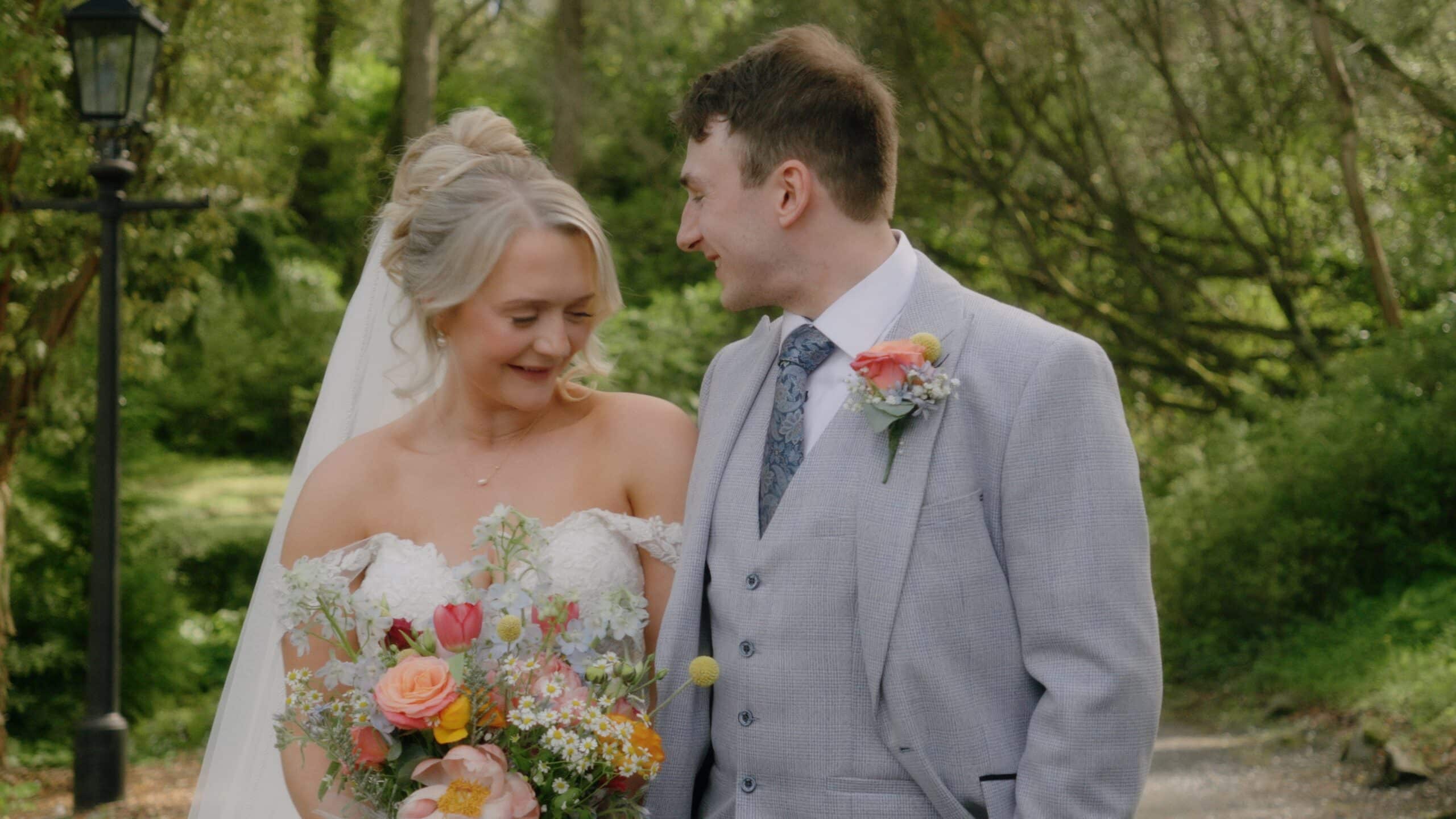 Bride and groom smiling in a garden setting, bride holding a colorful bouquet, groom in a light gray suit with floral boutonniere.