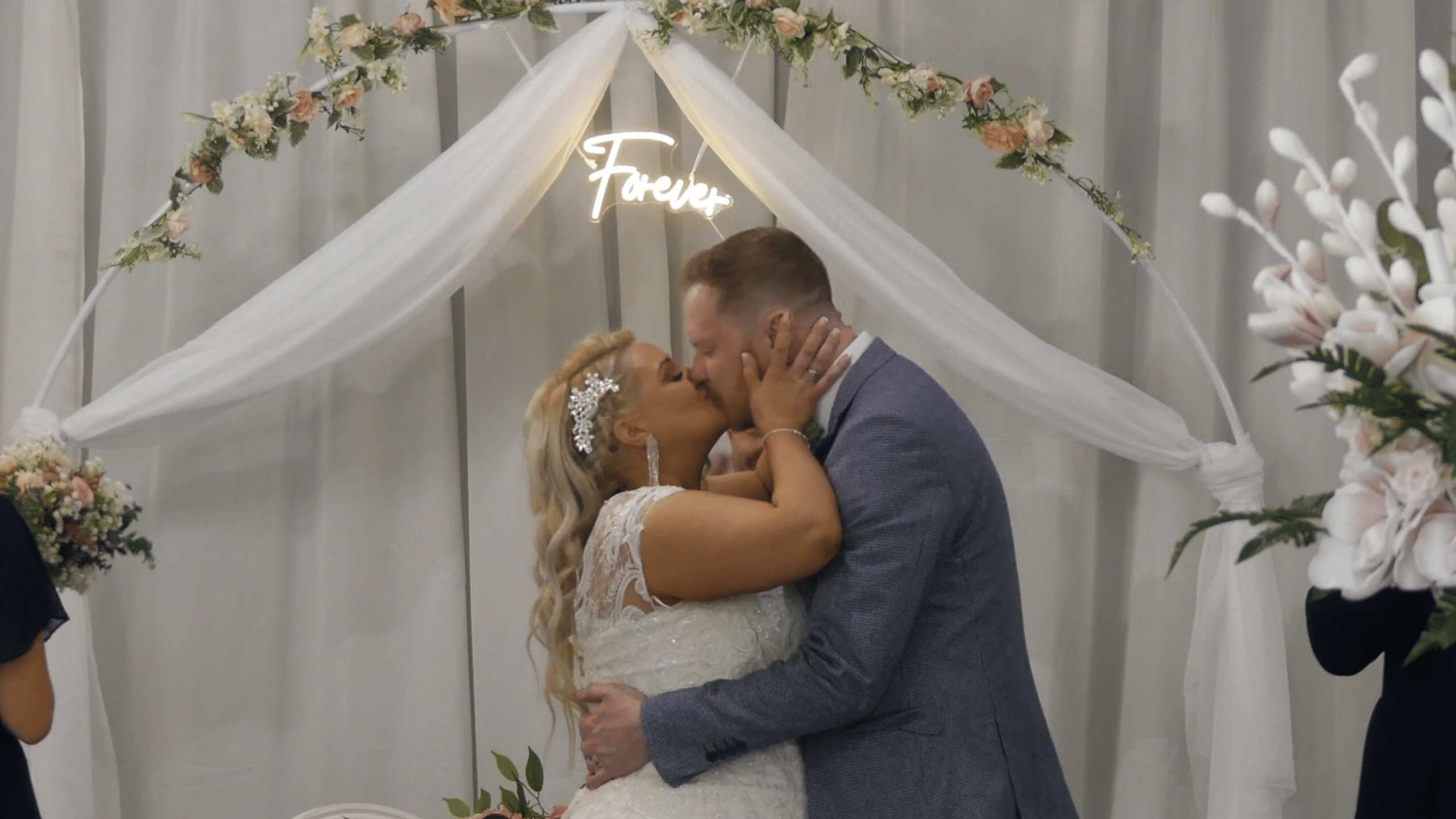 Bride and groom kissing under floral wedding arch with "Forever" sign.
