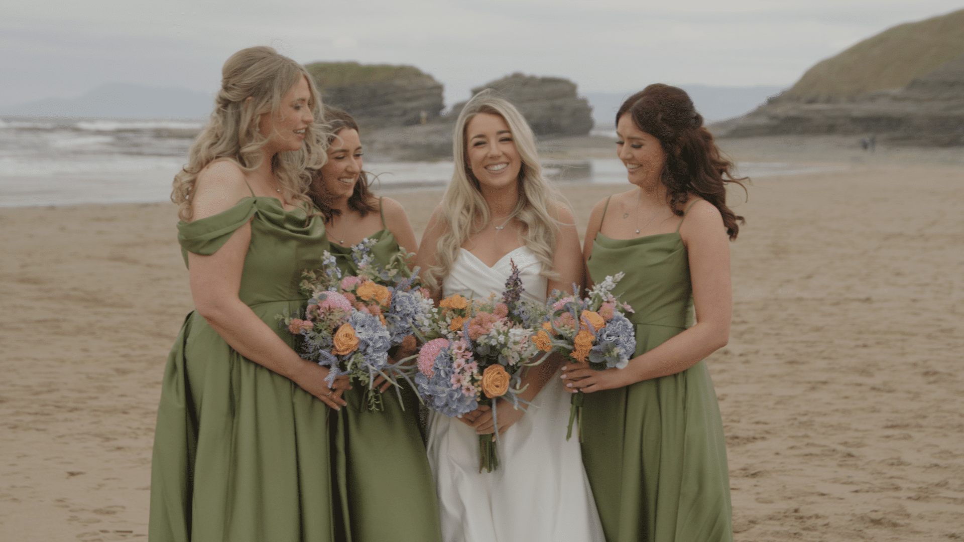 Bride and bridesmaids in green dresses holding colorful bouquets on a beach.