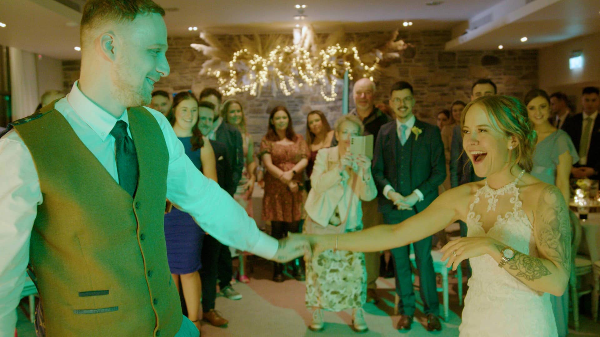 Bride and groom dancing at their wedding reception, surrounded by smiling guests and festive lights.