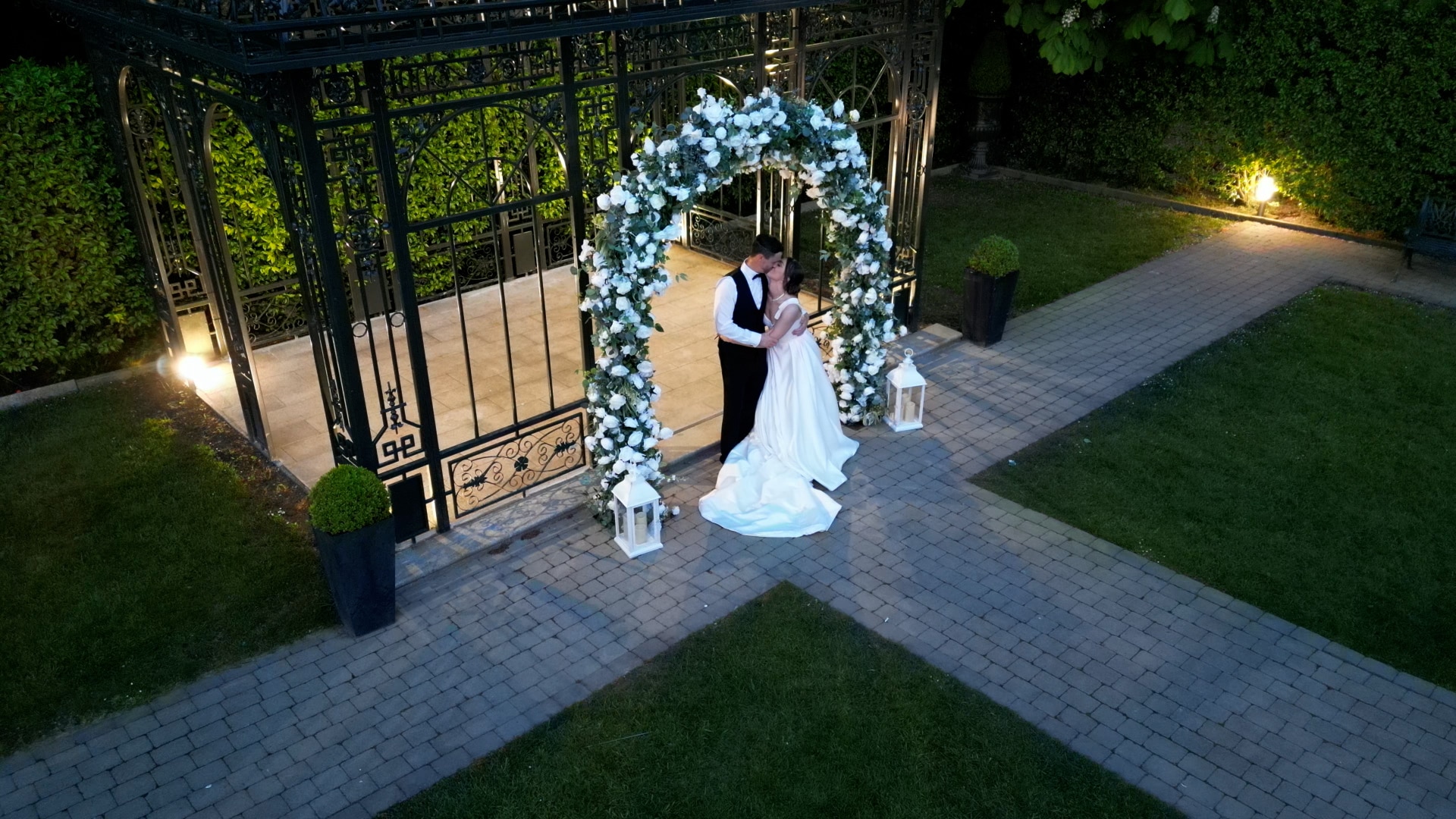 Bride and groom embracing under a floral arch at an outdoor venue in the evening.