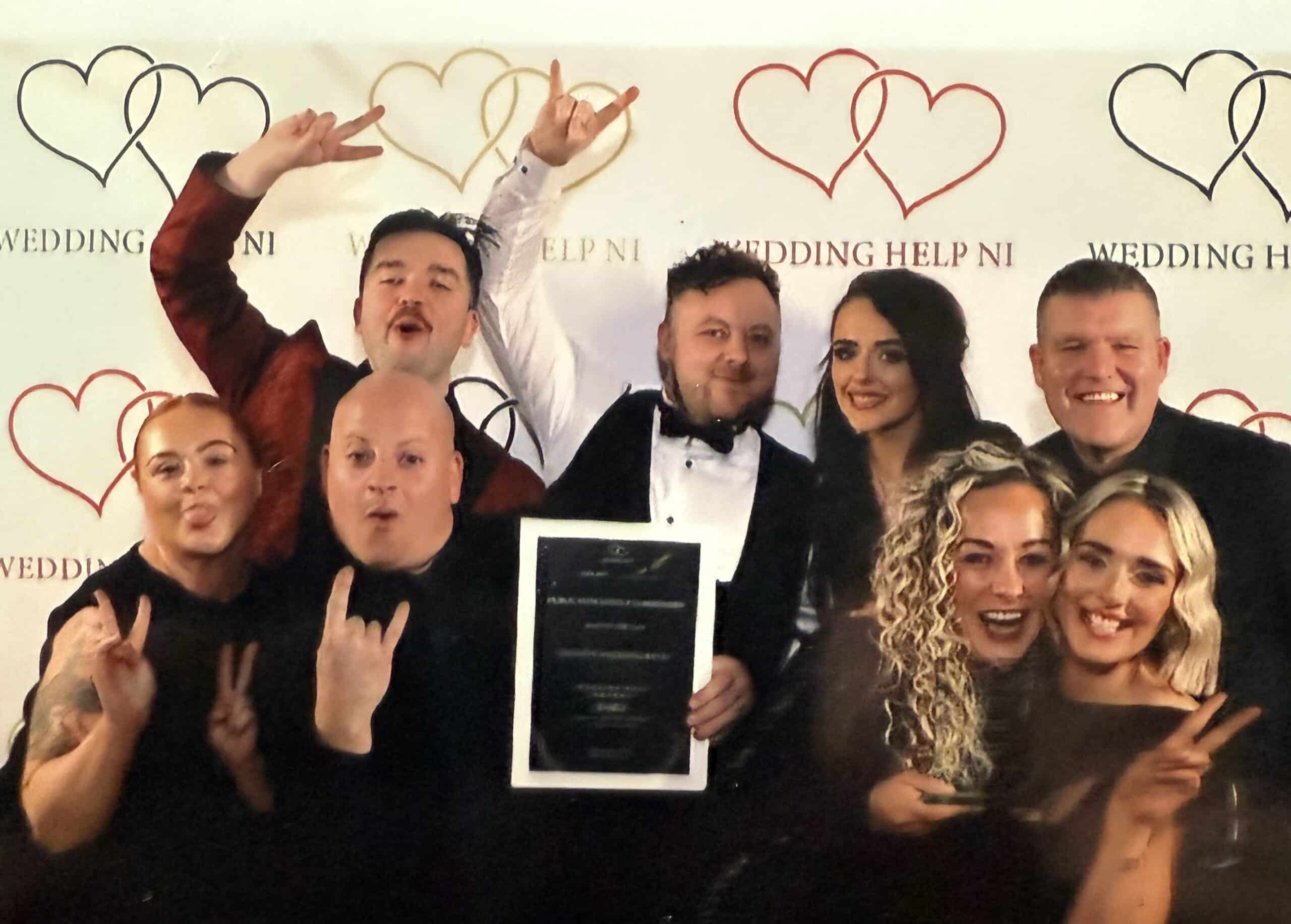 Group of people celebrating with a framed award, posing enthusiastically in front of a "Wedding Help NI" banner.