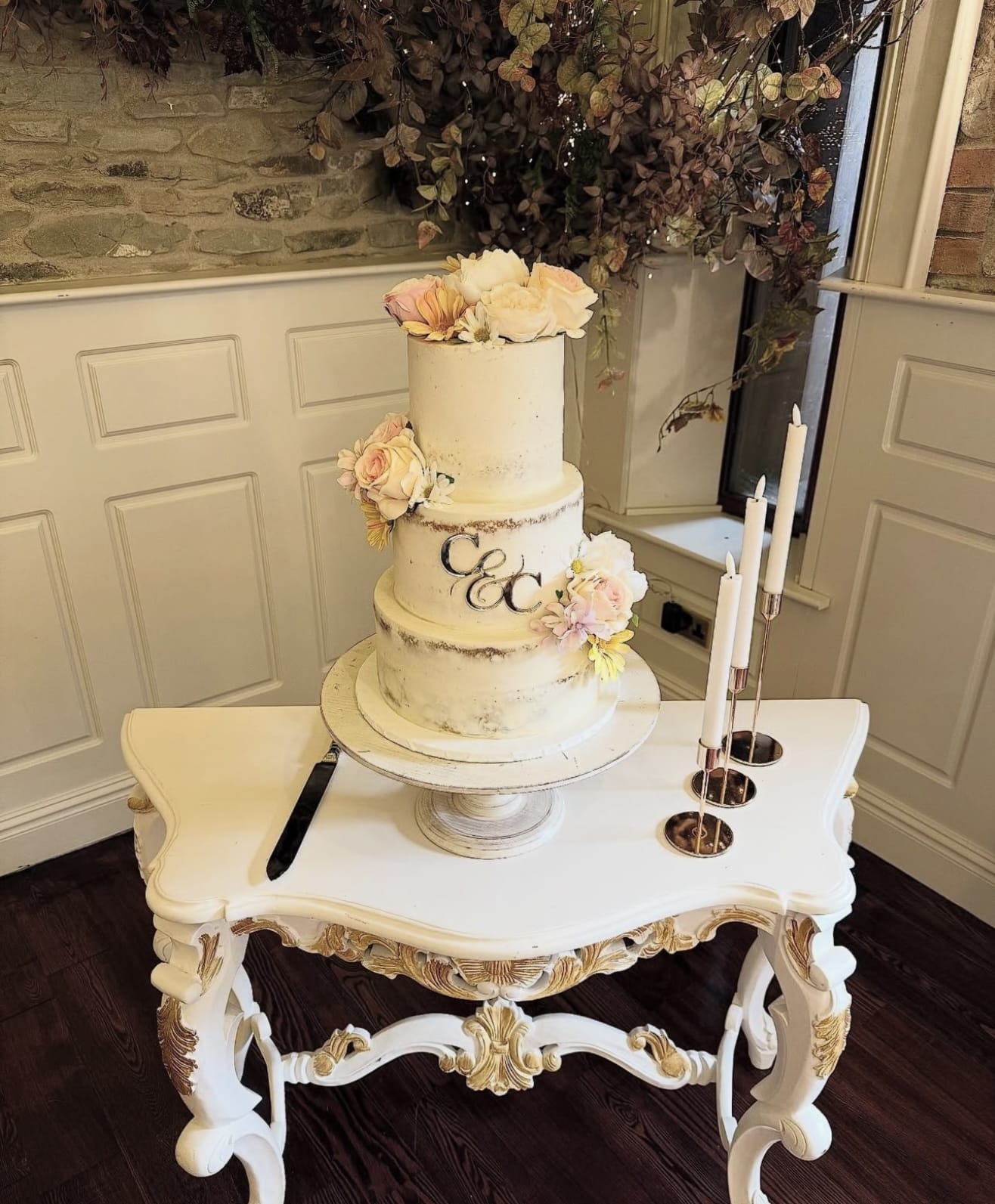 Three-tiered wedding cake with floral decorations on ornate white table with candlesticks.