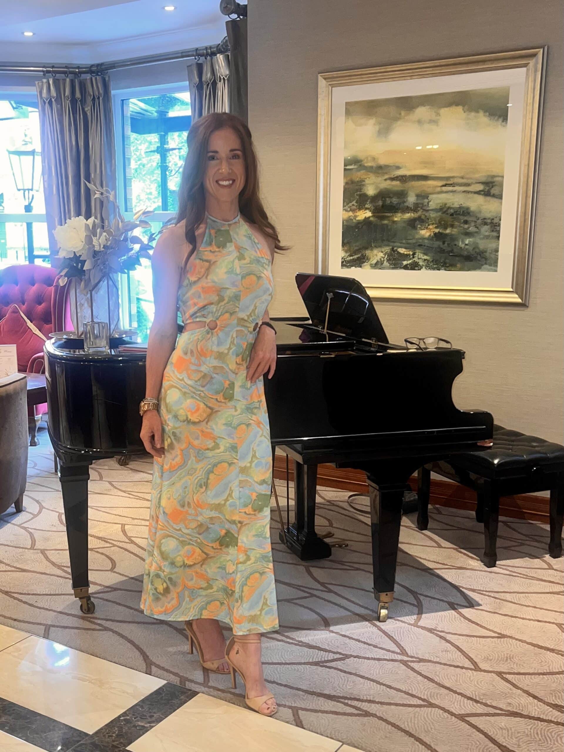 Woman in a colorful dress poses by a piano in a well-decorated room with art and flowers.