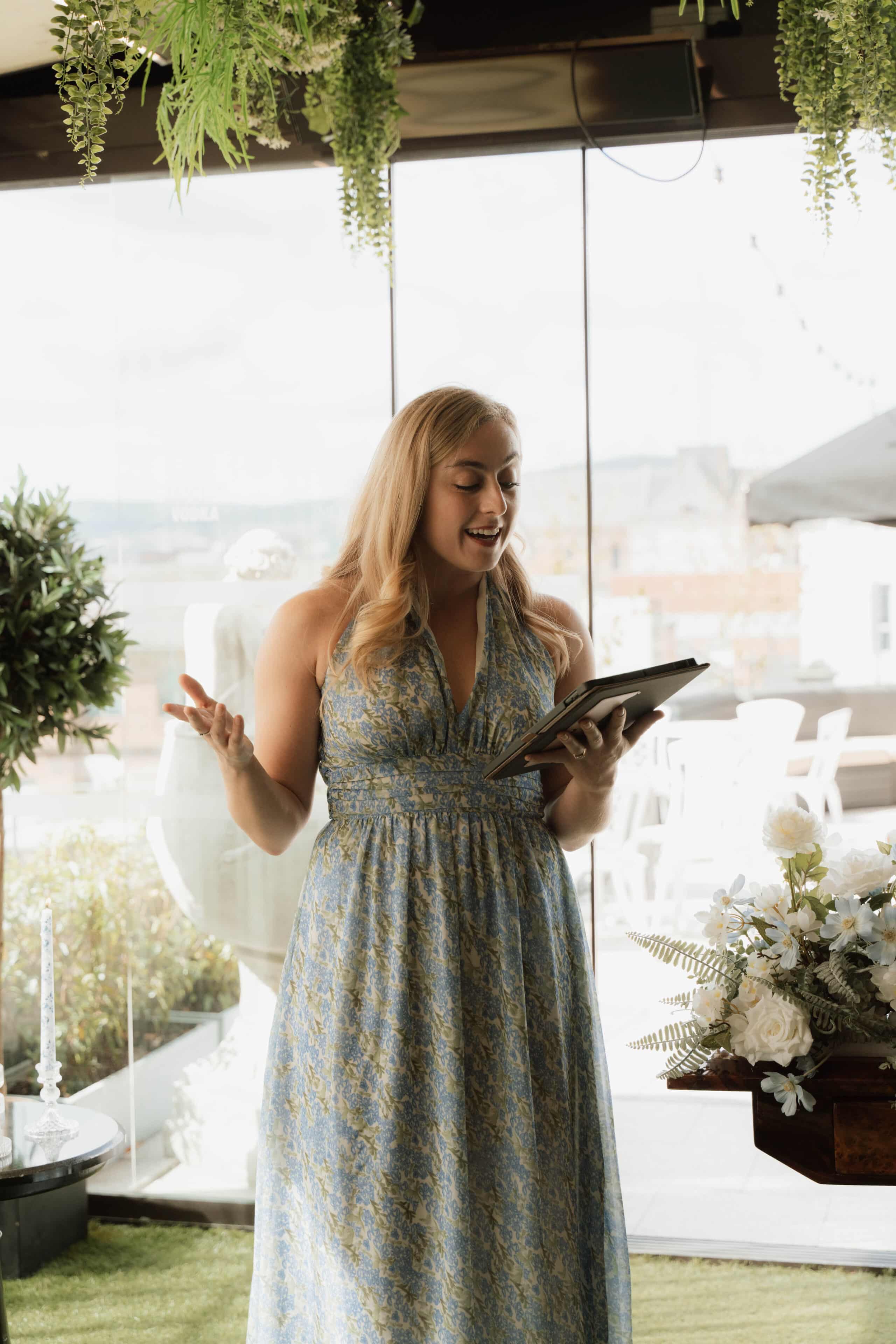 Woman in a blue floral dress reading from a tablet during an indoor event with greenery and flowers.