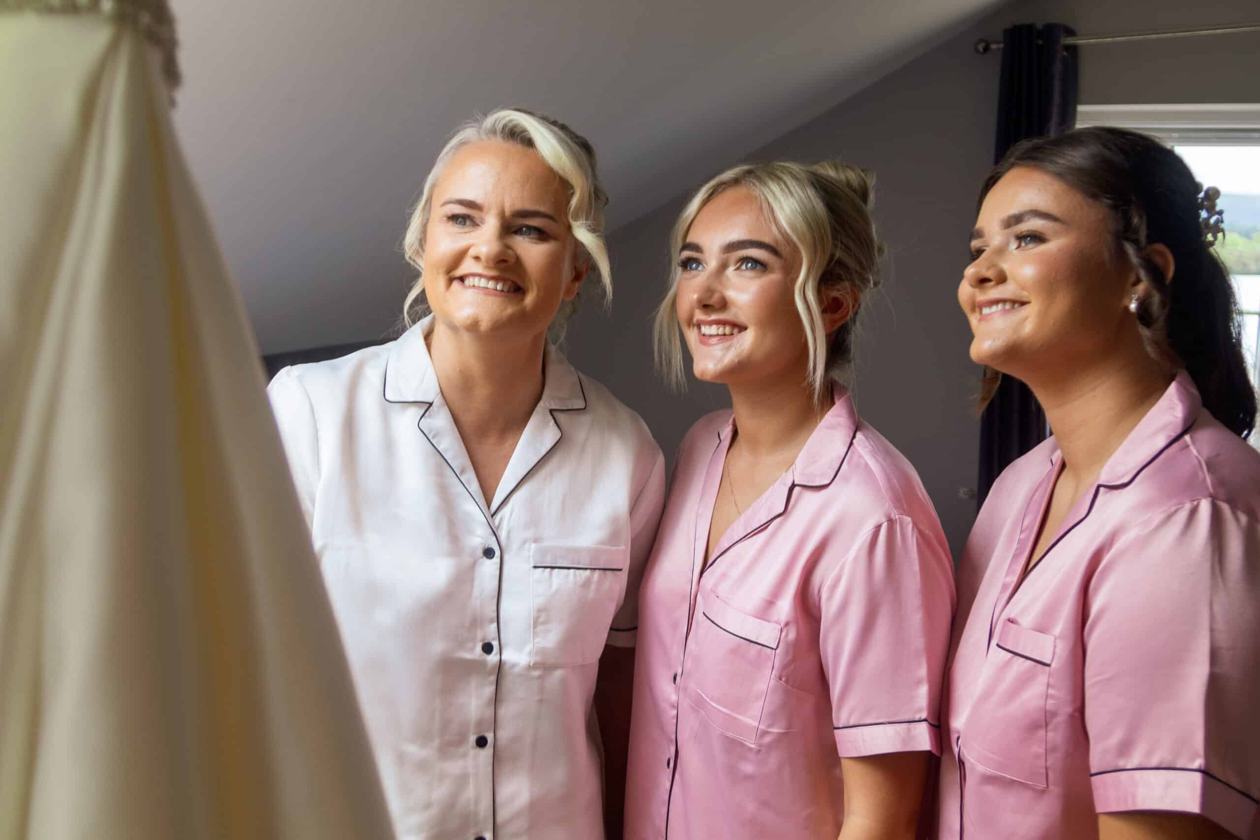 Three women in matching pajamas smiling at a wedding dress in a bright room.