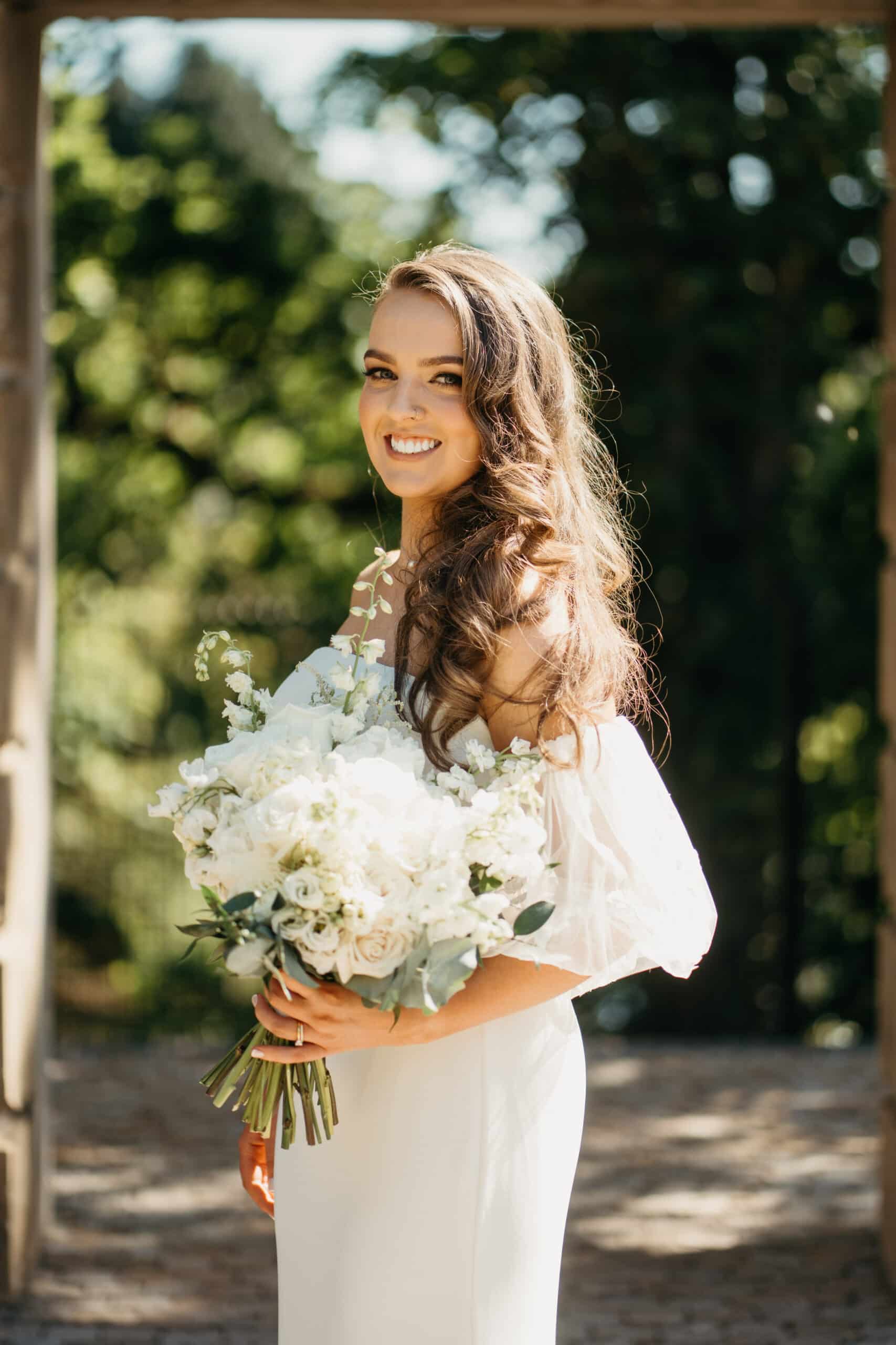 Bride smiling, holding a bouquet of white flowers outdoors.