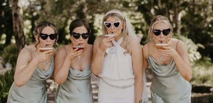 Bride and bridesmaids with heart sunglasses sipping cocktails outdoors.