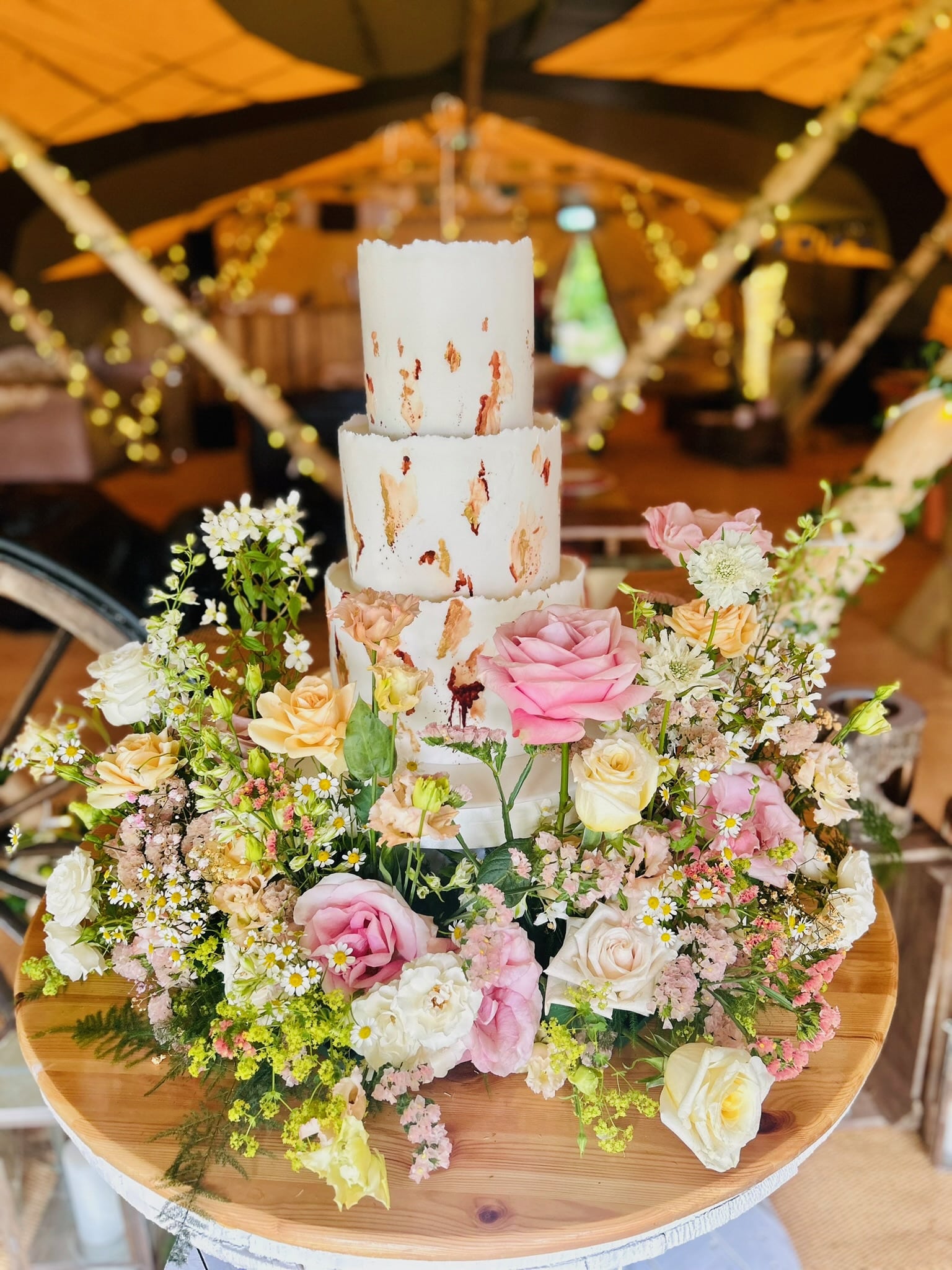 Three-tier cake with rustic icing adornments, surrounded by colorful flower arrangements on a wooden table.