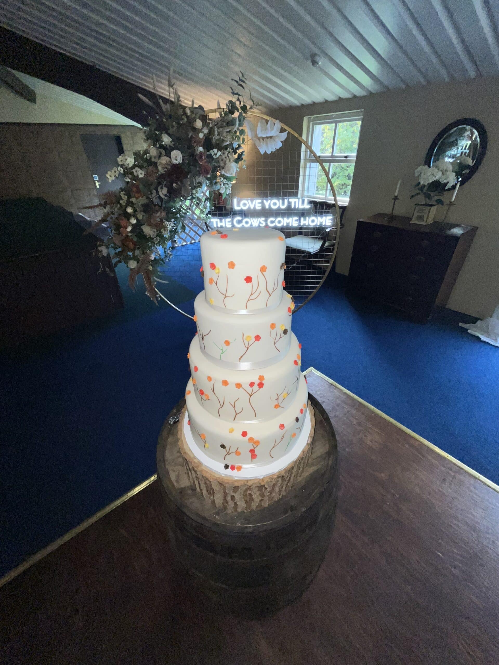 Four-tier wedding cake with floral design, on a wooden barrel, under a neon sign reading "Love you till the cows come home."