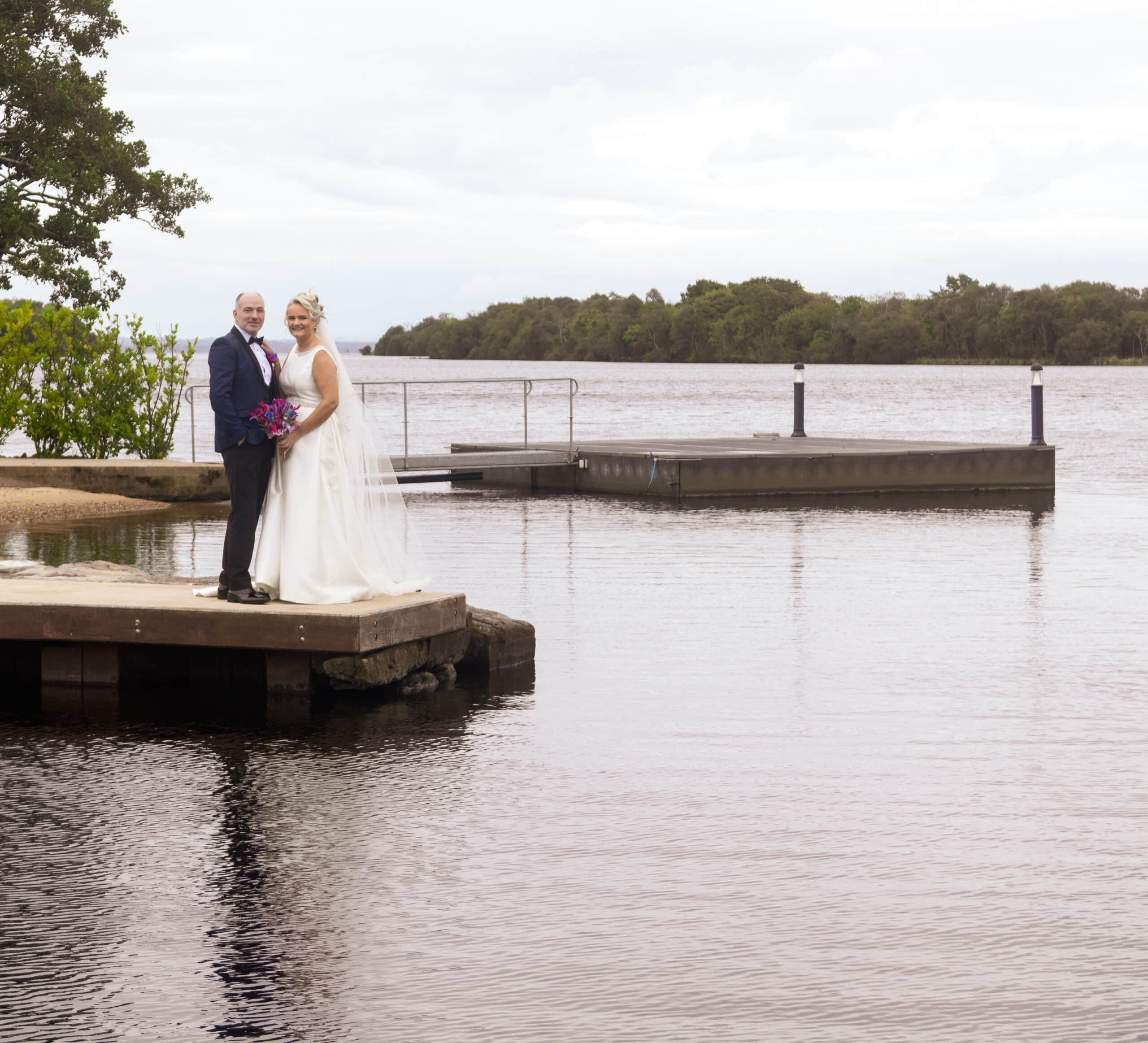 Bride and groom standing on a riverside dock, surrounded by tranquil water and greenery.