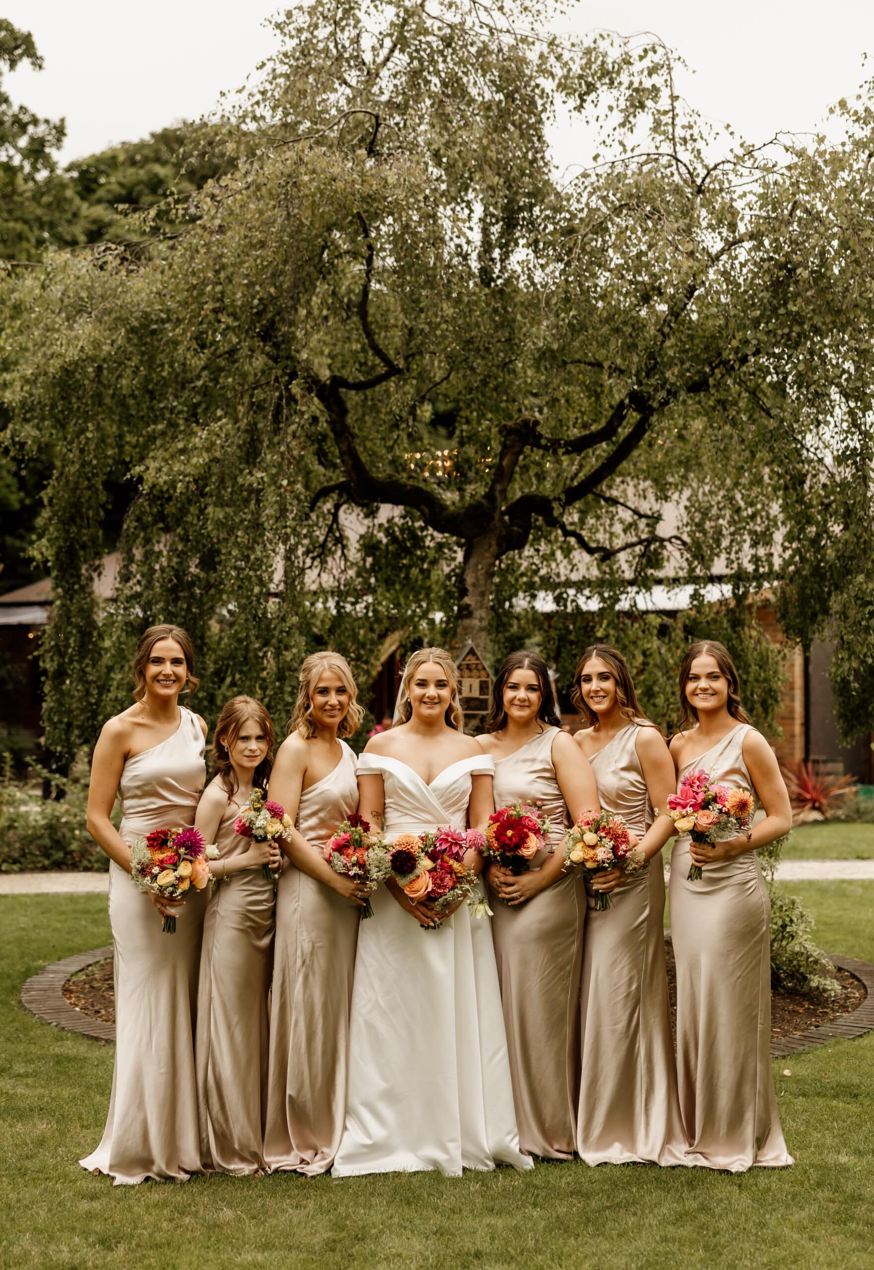 Bride and bridesmaids in elegant dresses holding flowers, standing in a garden setting.