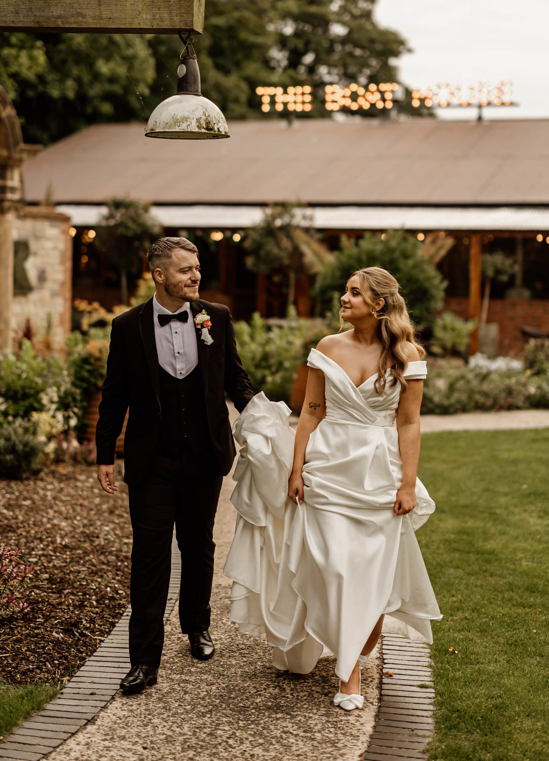 Bride and groom walking outside venue, groom in a black tuxedo, bride in a white gown, lush greenery in the background.