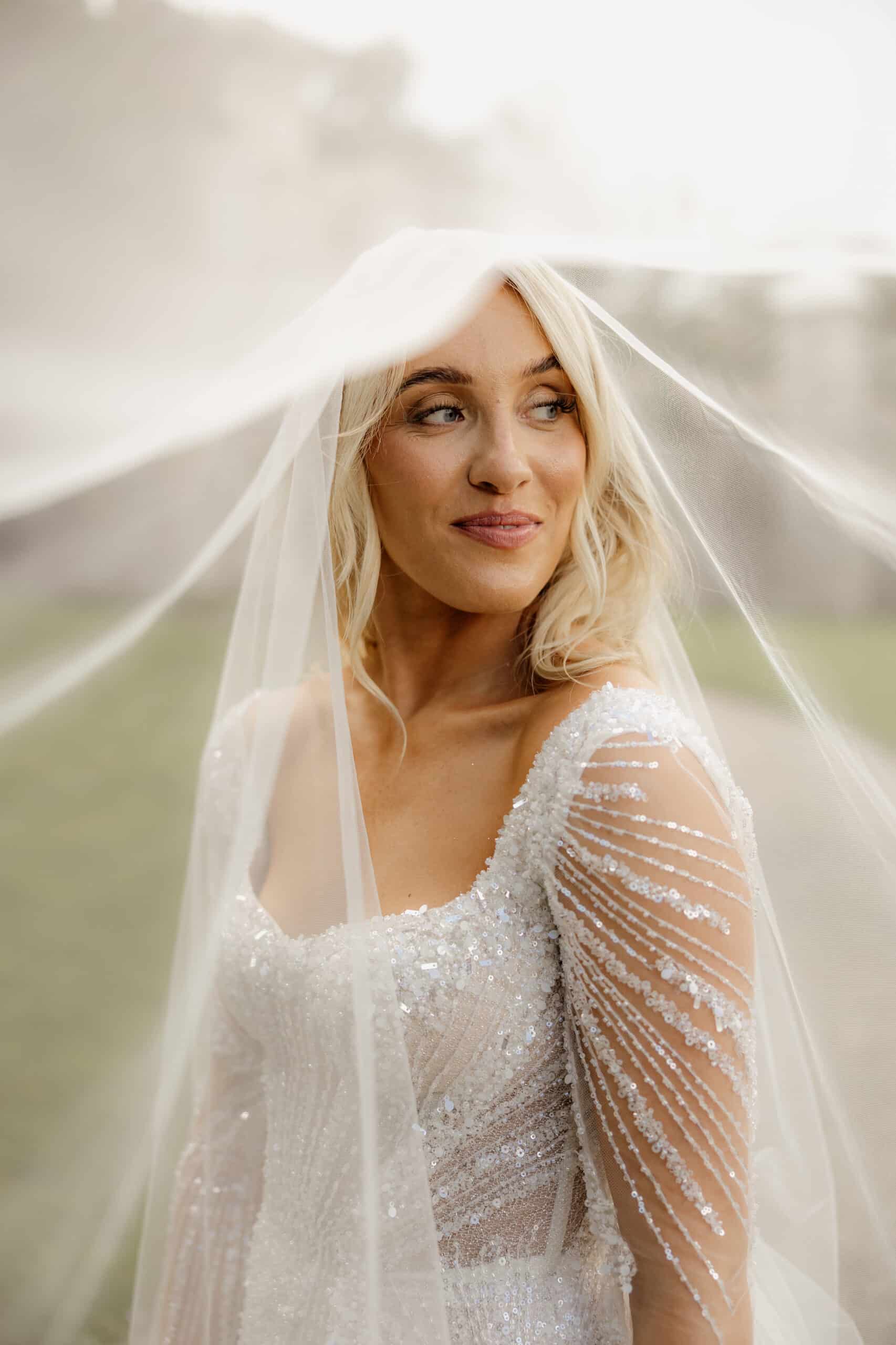 Bride in a sparkling wedding dress and veil smiles softly, looking away from the camera.