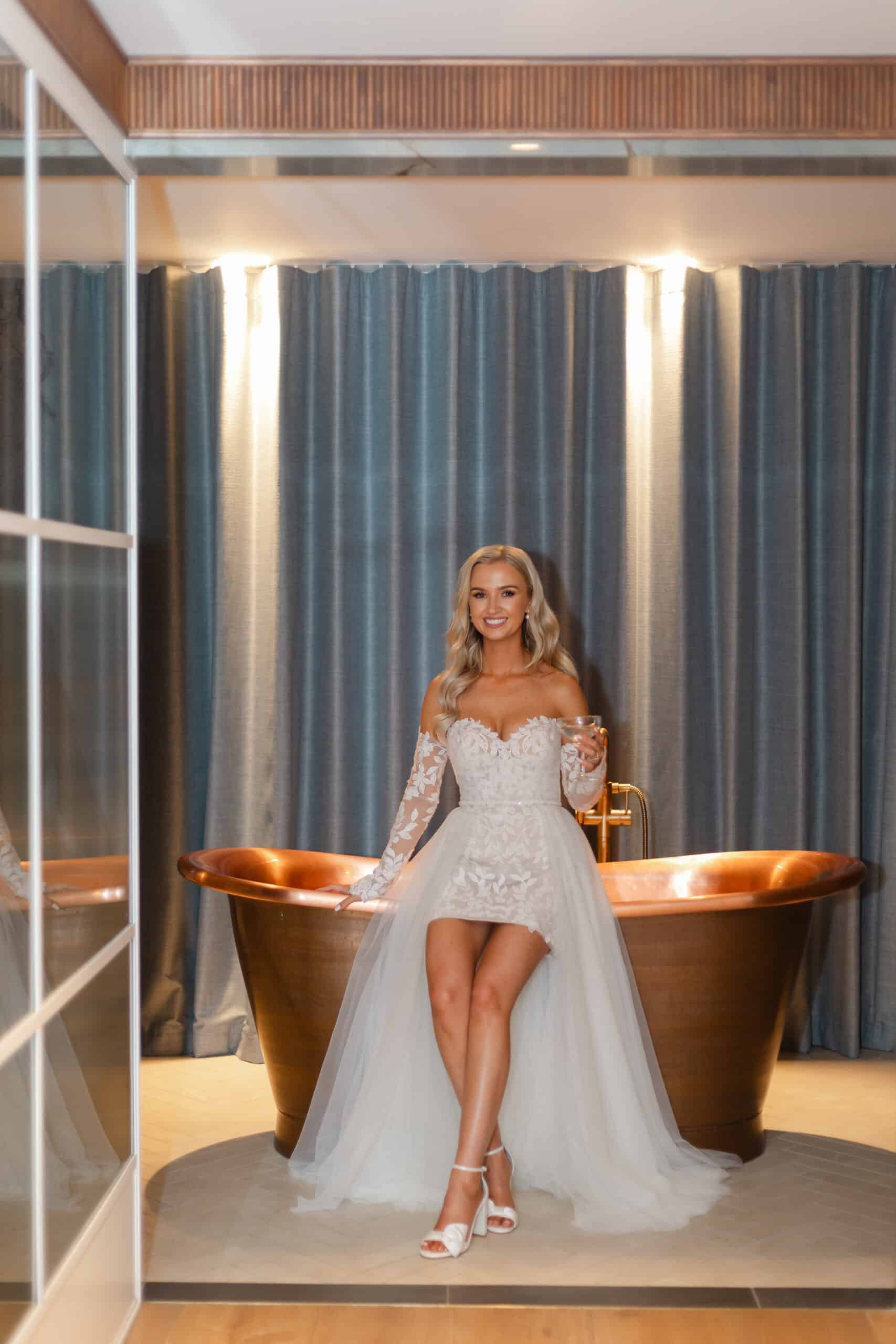 Bride in a lace wedding dress poses with a drink in front of a copper bathtub.