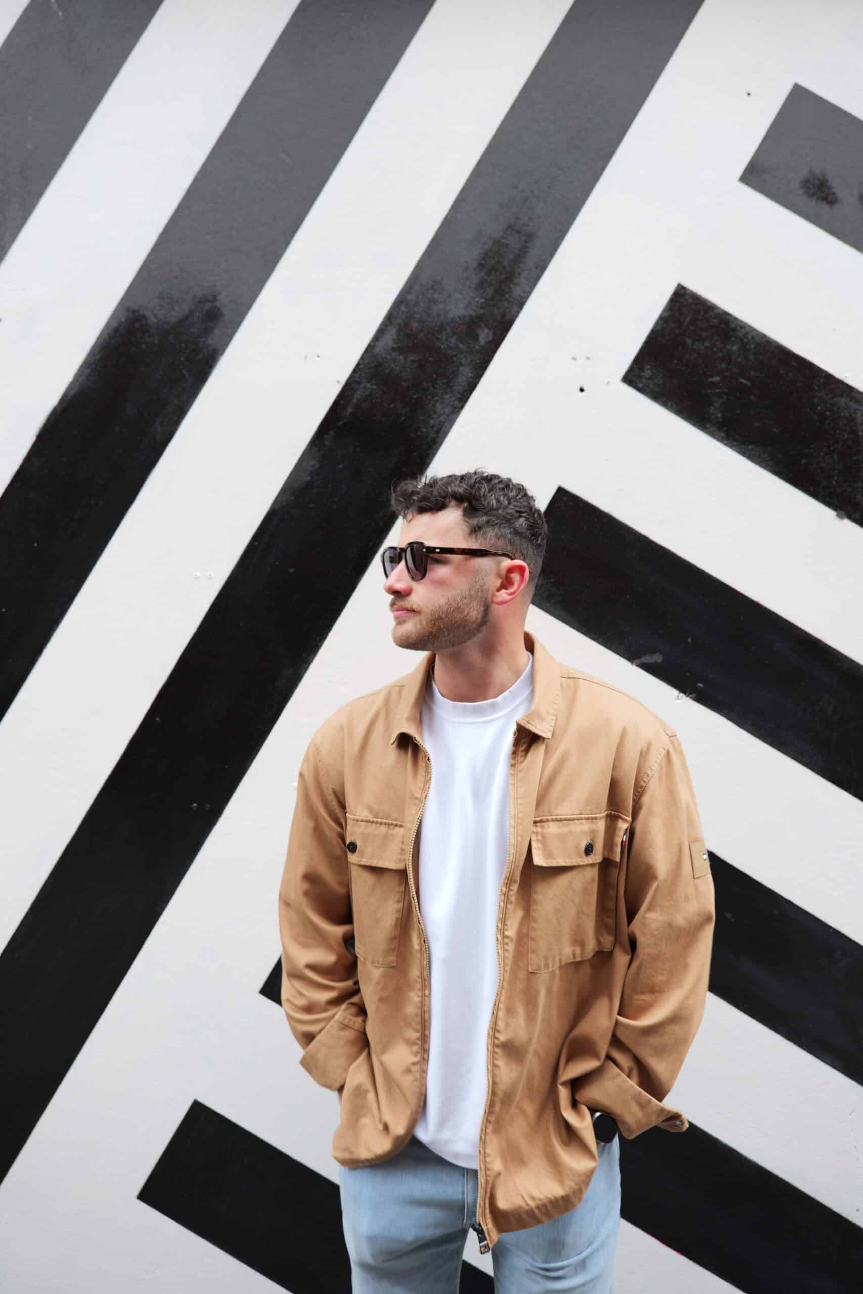 Man in tan jacket and sunglasses standing against a geometric black and white striped wall.